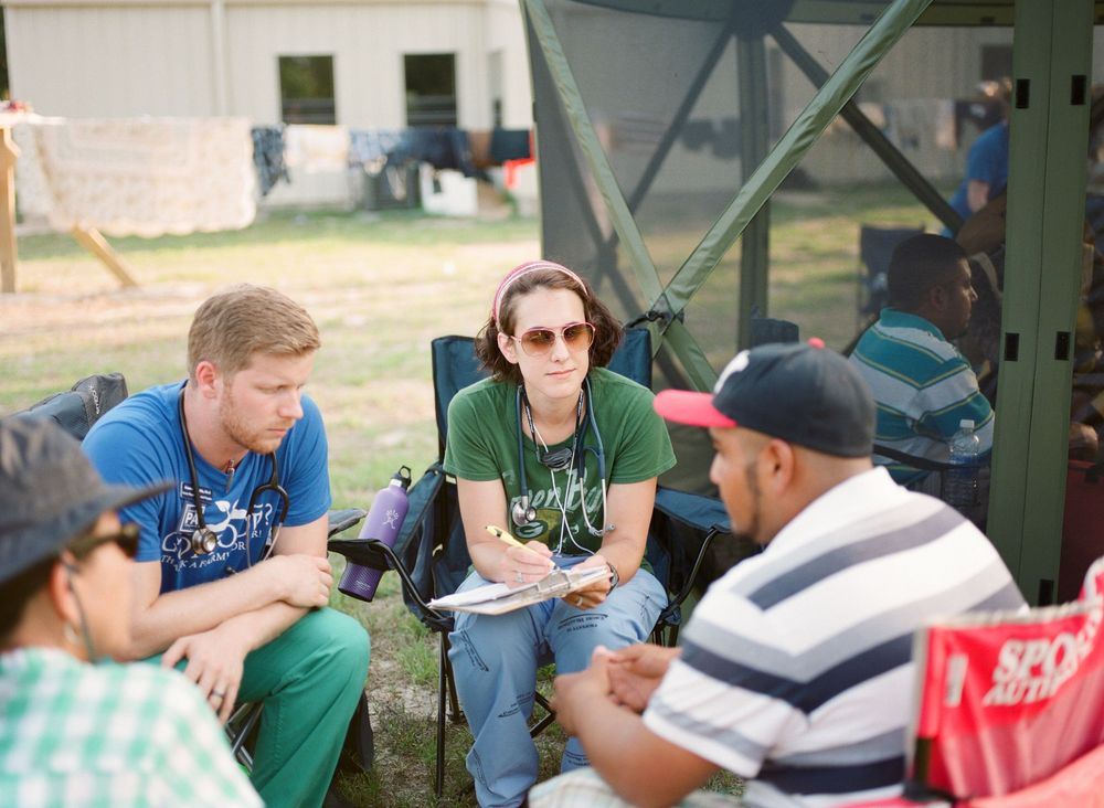 Students sit at outdoor tables engaged in group study and discussion activities on a sunny campus day.