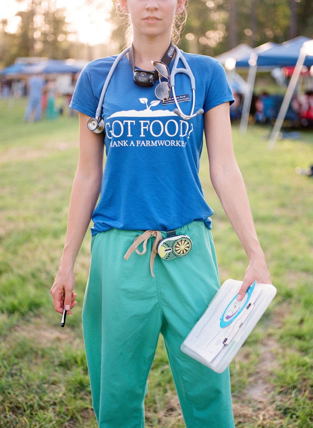 Person wearing blue t-shirt and green overalls stands in outdoor field holding white container in golden sunlight.