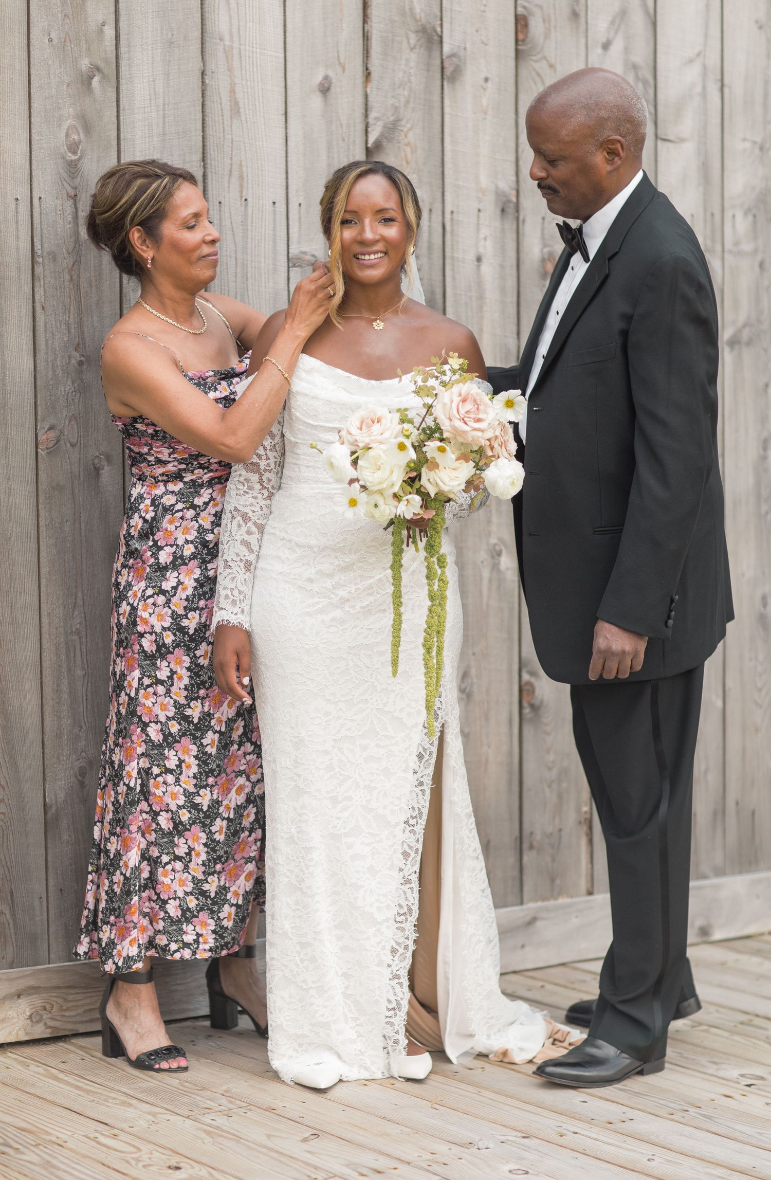 A bride in a white lace gown holds a cream bouquet while standing with two others against a wooden fence background.