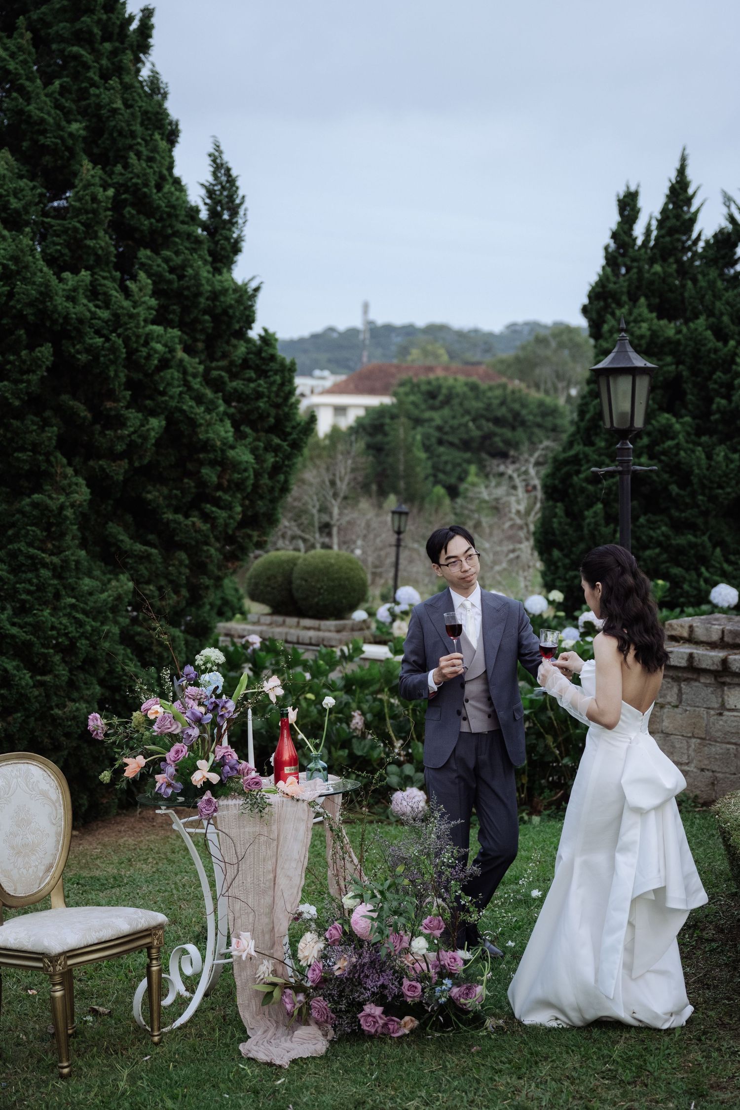 Groom in gray suit and bride in white gown share wine near a floral cart in a garden.