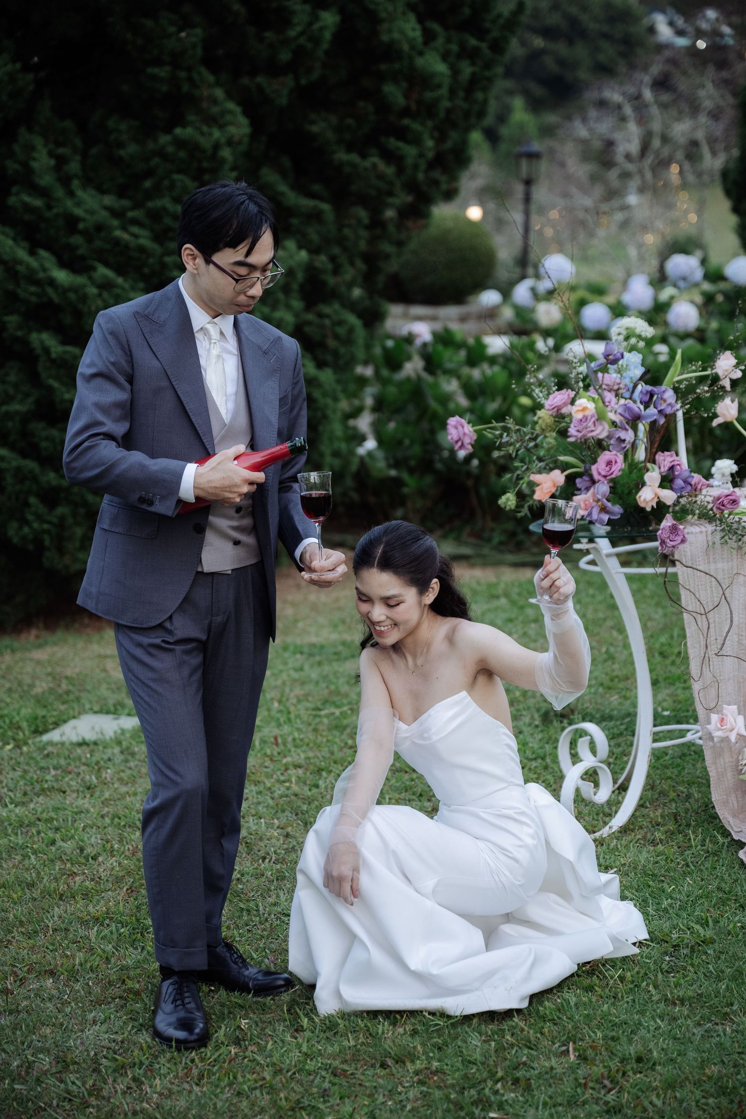 Groom reads vows while bride kneels laughing, both holding wine glasses in an outdoor garden setting.