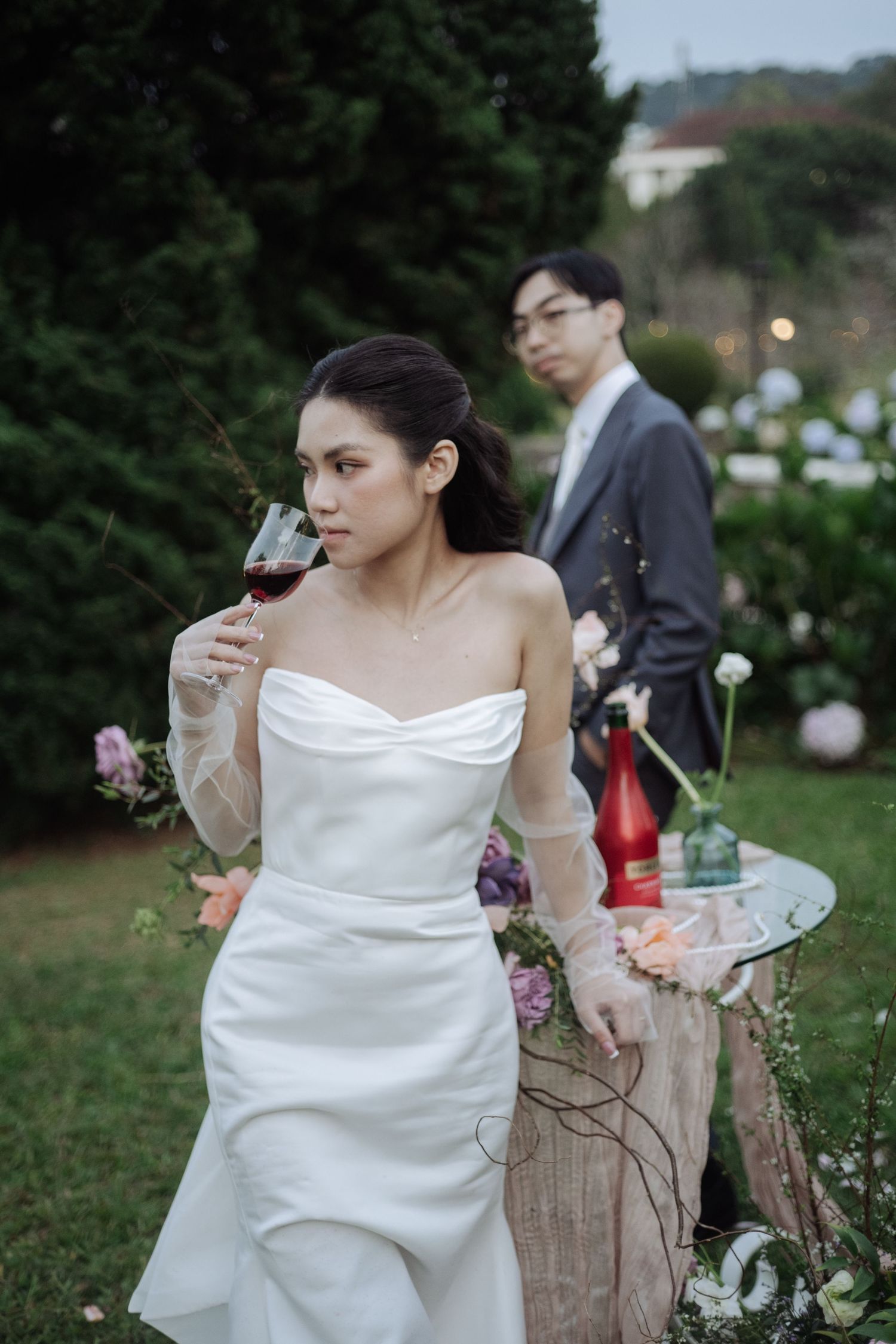 Elegant bride in strapless white gown sips red wine in a lush garden at dusk.