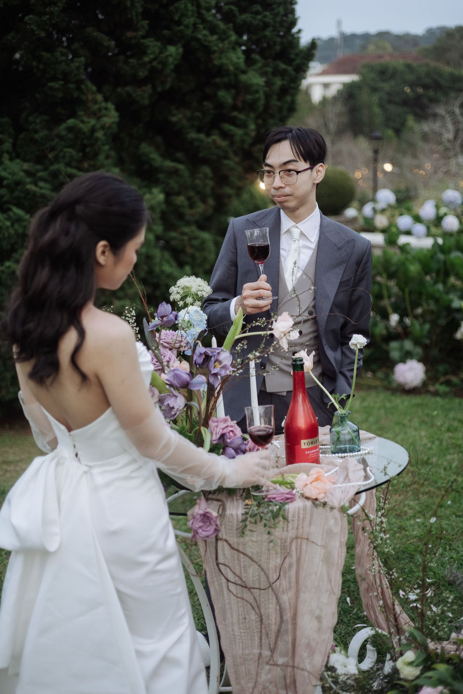 Bride and groom share a toast with red wine beside a floral display in an evening garden.