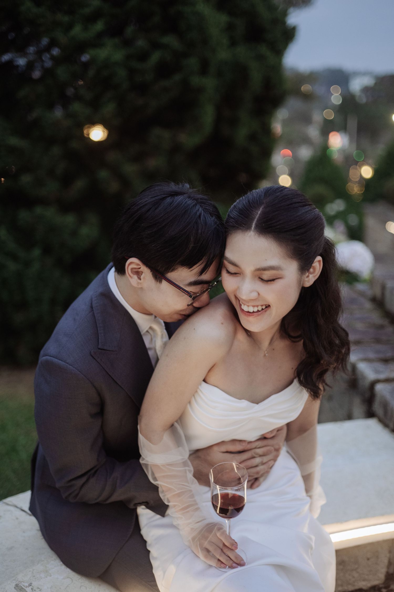 Groom tenderly kisses brides shoulder as she laughs joyfully, holding a wine glass at twilight.