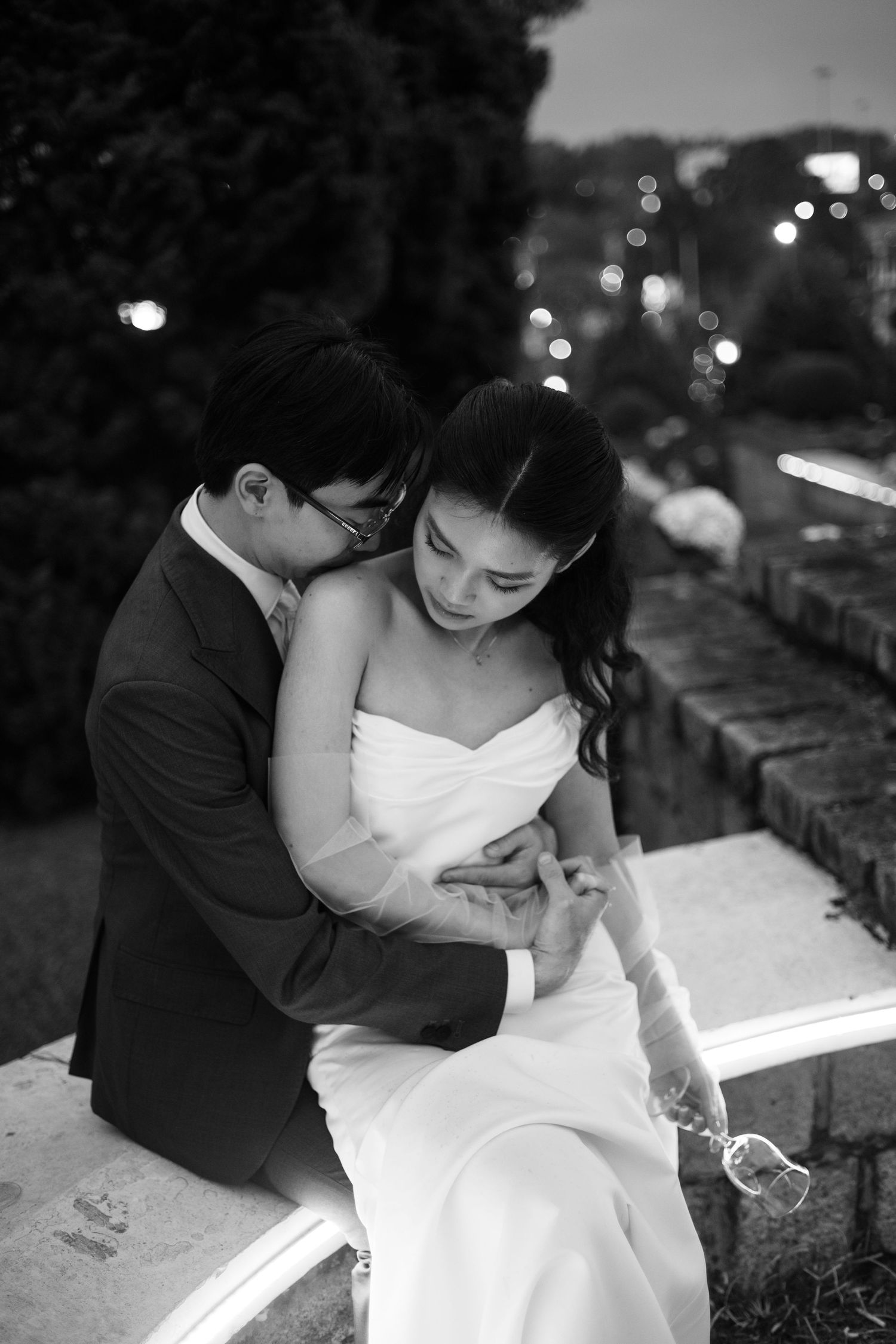 Black and white romantic portrait of groom embracing bride while she holds a wine glass at dusk.