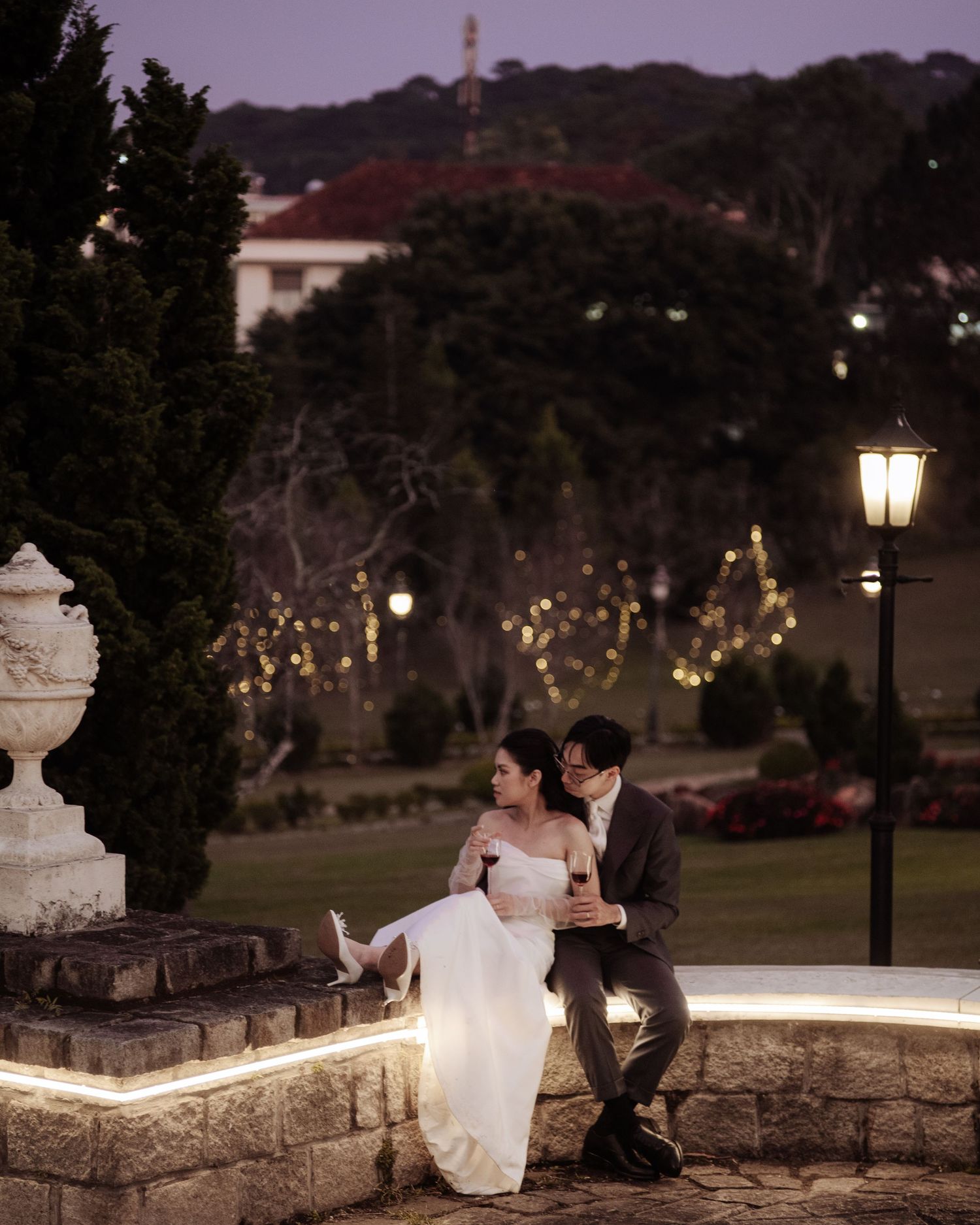 Couple in wedding attire sitting on stone wall at dusk, surrounded by garden lights and lush greenery.