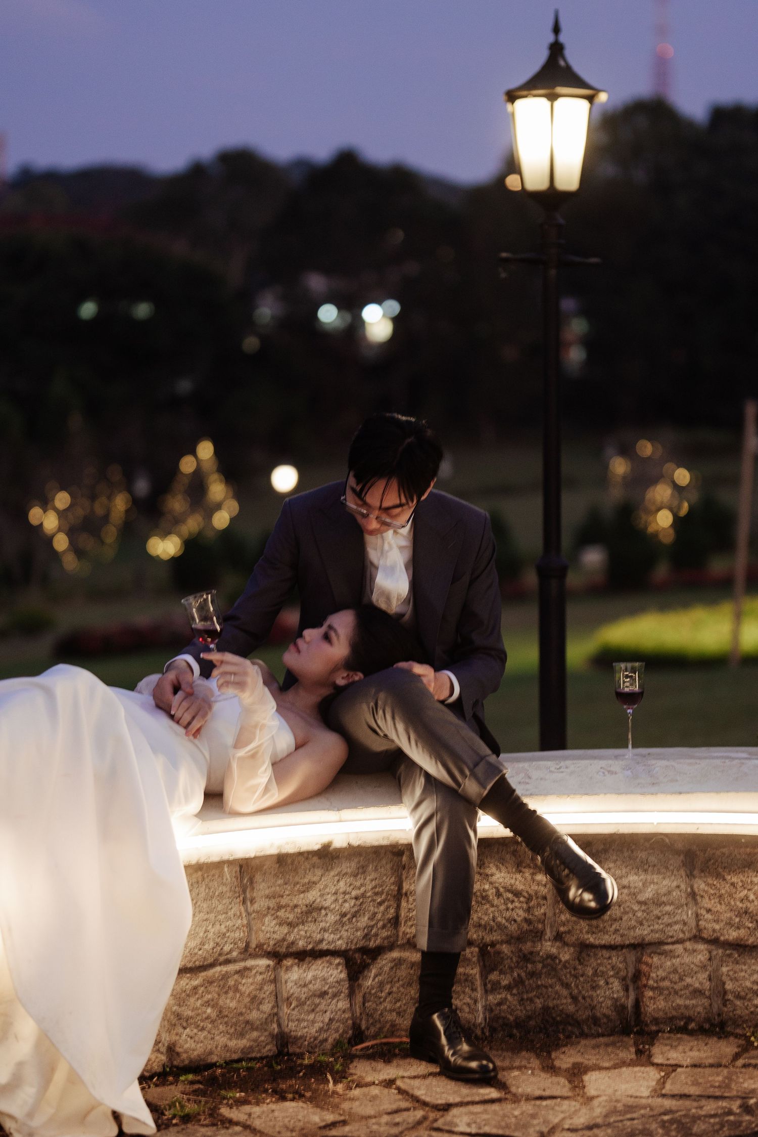 Bride and groom sharing romantic moment on illuminated stone ledge at twilight, holding wine glasses.