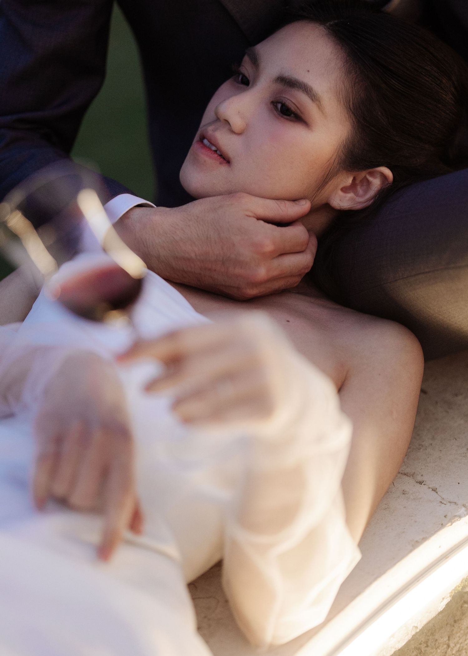 Close-up of bride in white dress lying down, groom tenderly holding her face in evening light.