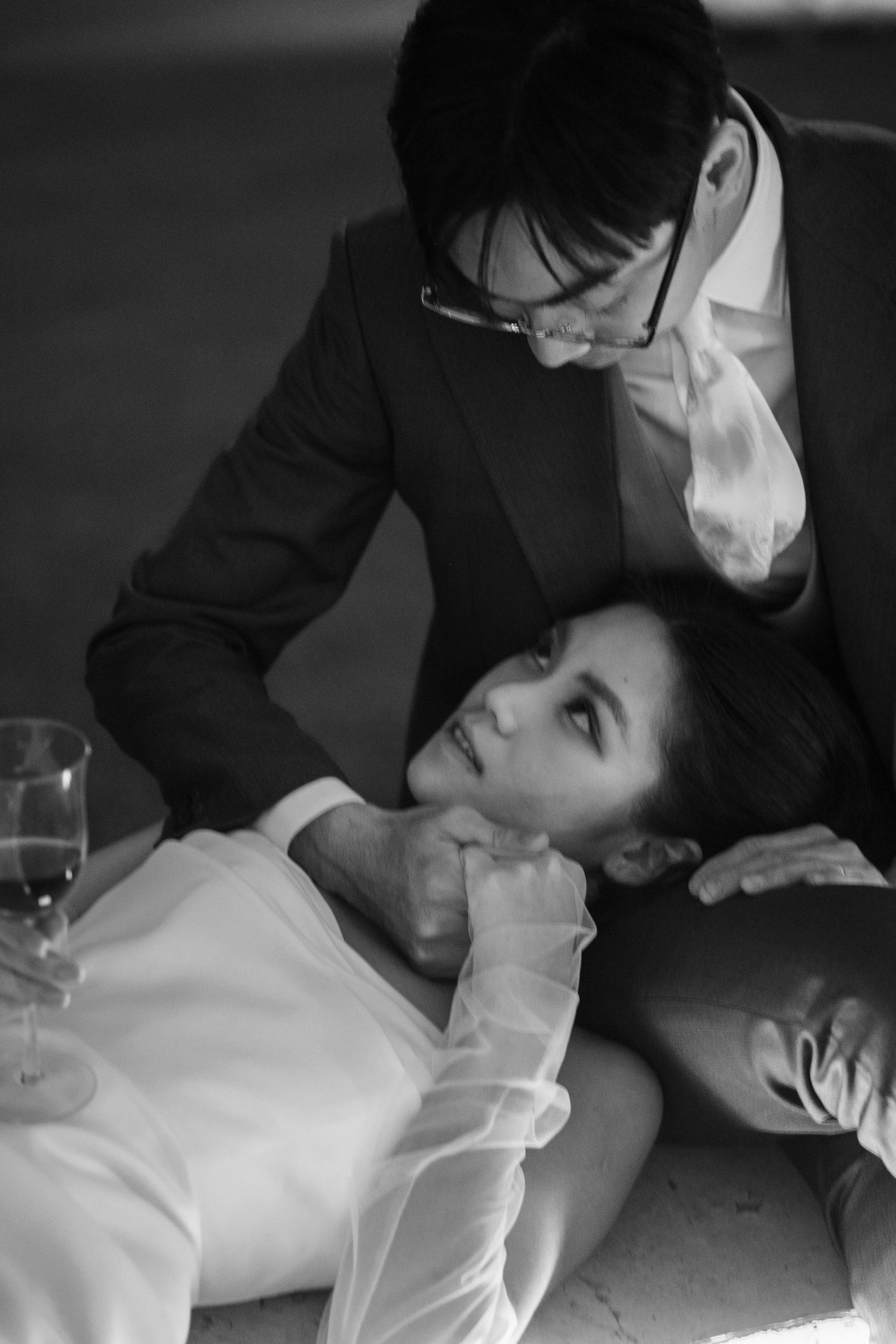 Black and white photo of groom gazing at bride lying across his lap near a wine glass.