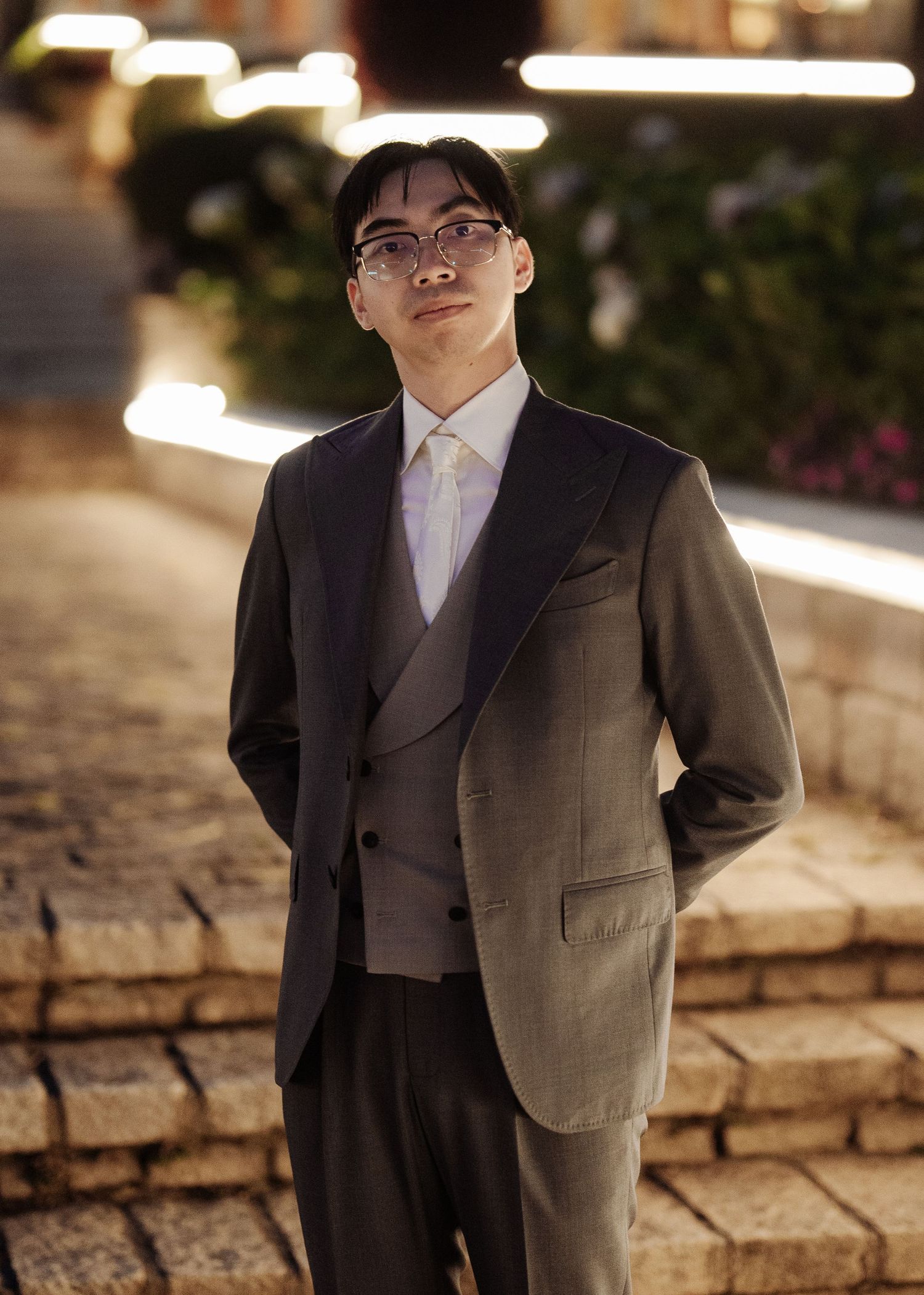 Dapper groom in three-piece suit and glasses standing on stone steps in evening garden setting.