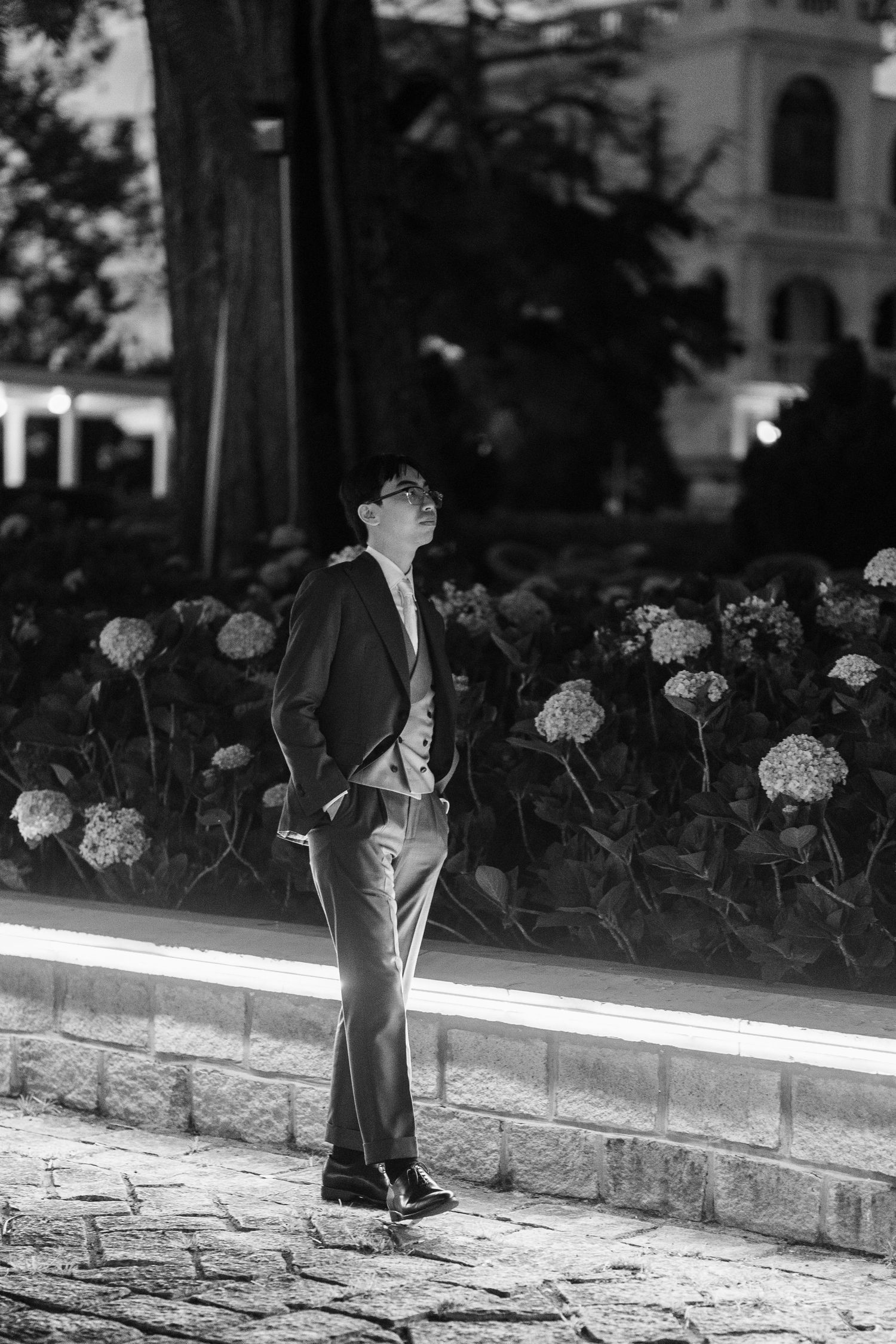 Groom in dark suit looking upward beside flower bed at night in black and white portrait.