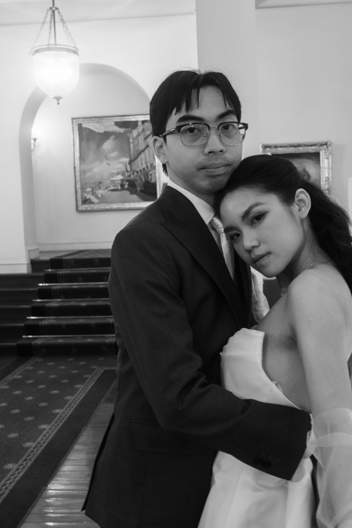 A black and white portrait of a couple in wedding attire embracing in an elegant indoor staircase setting.