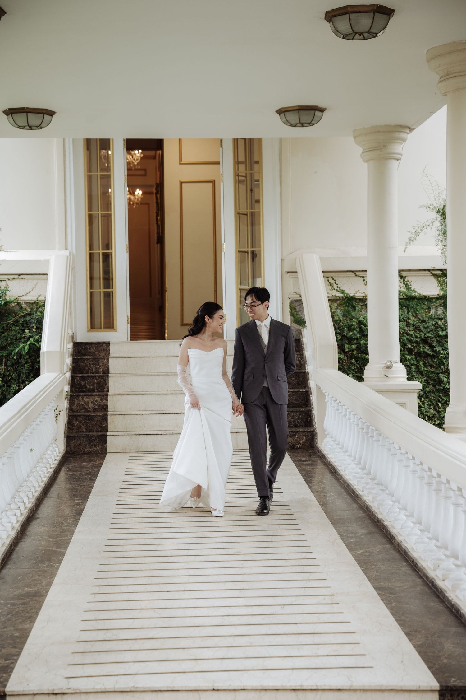 Elegant bride and groom walk hand in hand along a white columned veranda of a grand estate.