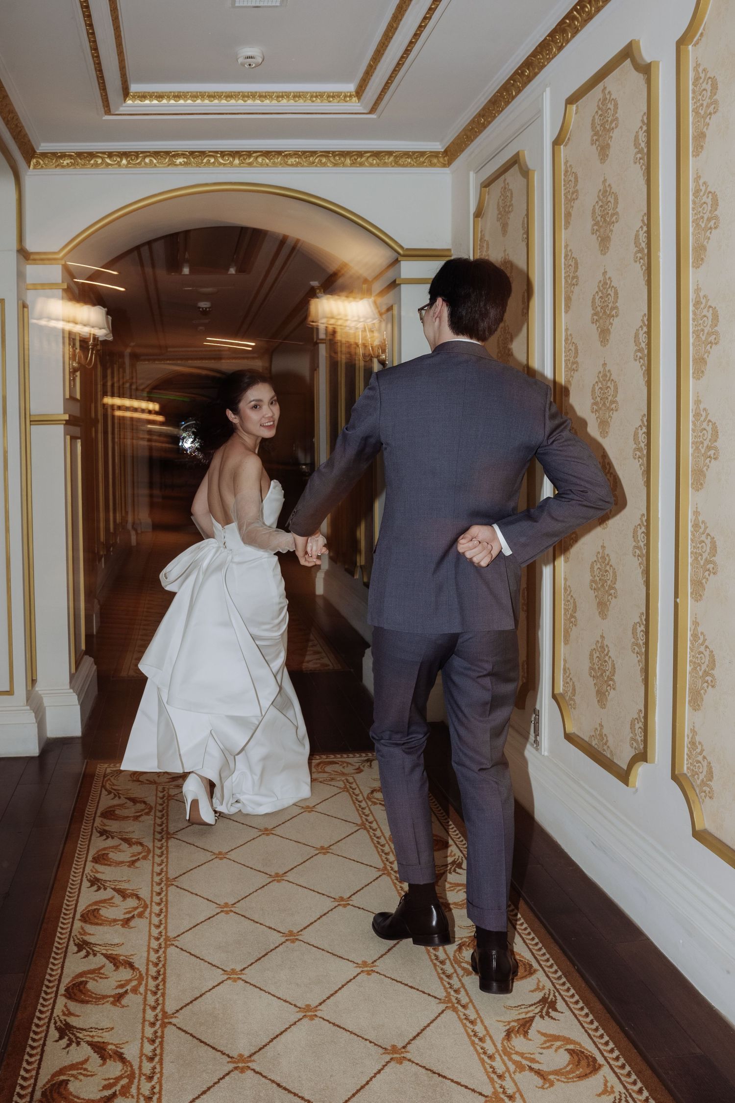 Bride in backless white gown leads groom through an ornate golden hallway of a luxury hotel.