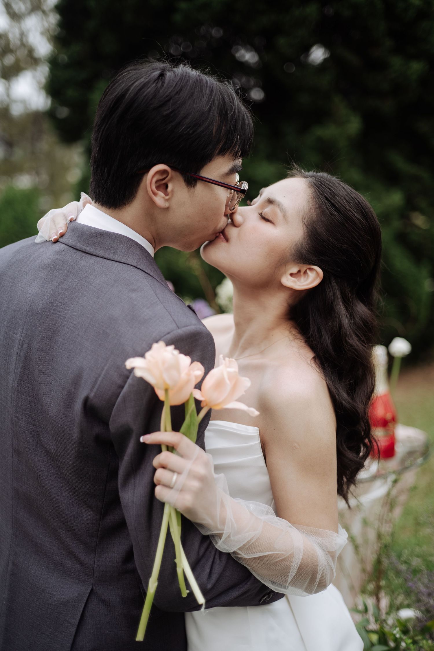 Bride and groom share a tender kiss outdoors while holding soft pink tulips in a garden.