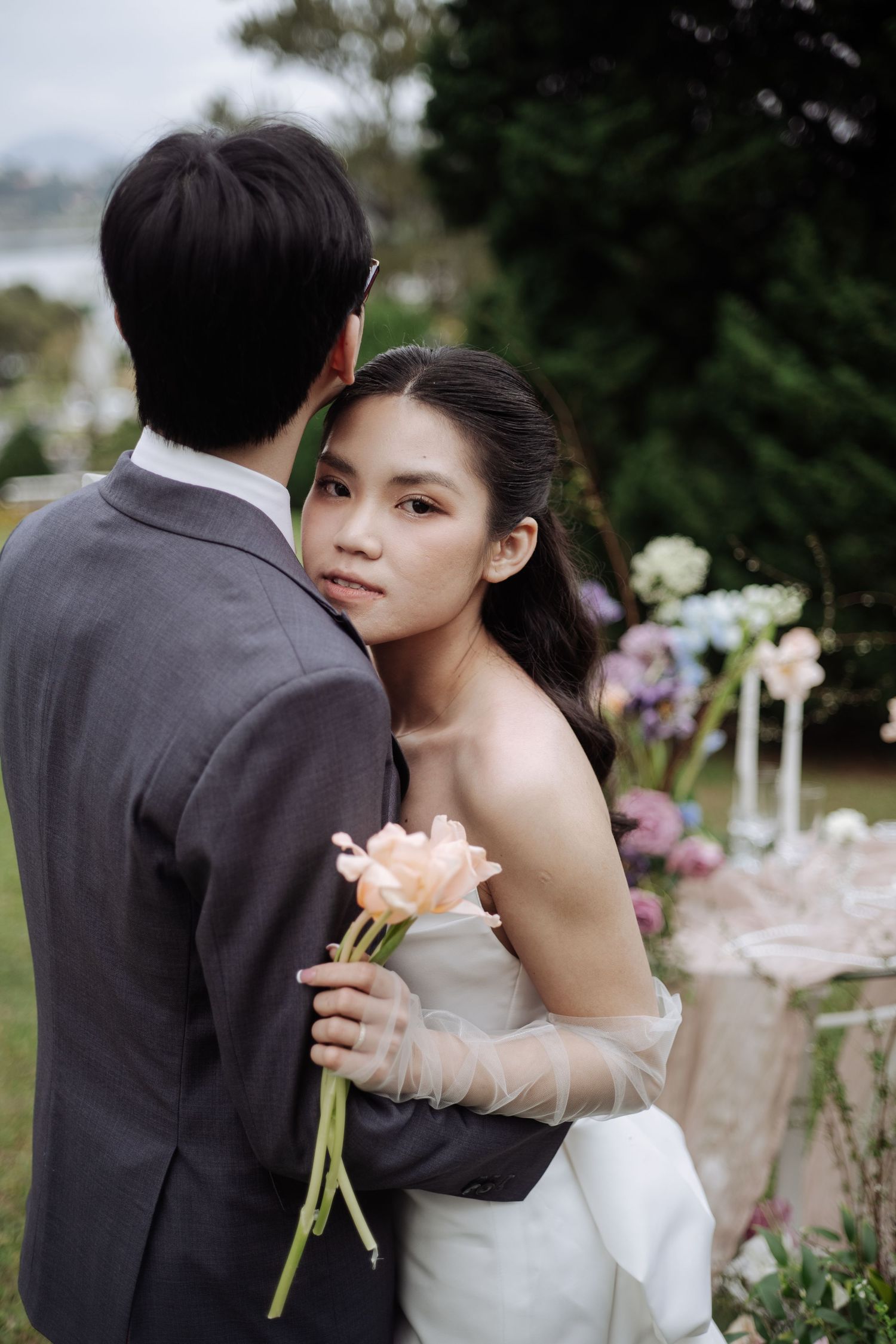 Bride in strapless white gown embraces groom while holding a peach rose at outdoor wedding.