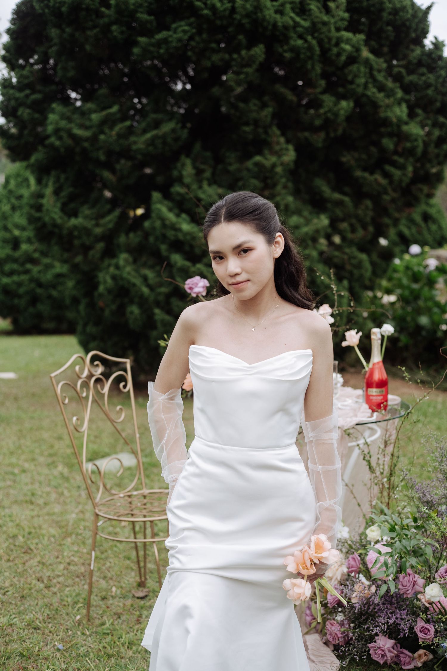 Elegant bride in strapless white satin gown poses in a lush garden with colorful floral decor.