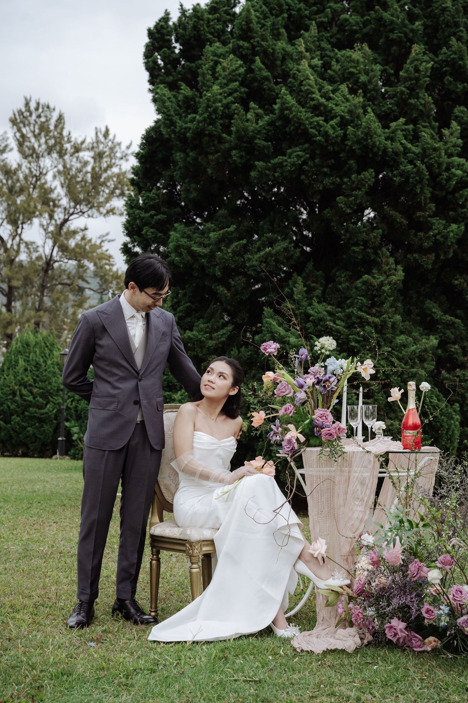 Groom gazes at seated bride beside an elegant floral arrangement in a garden wedding setup.