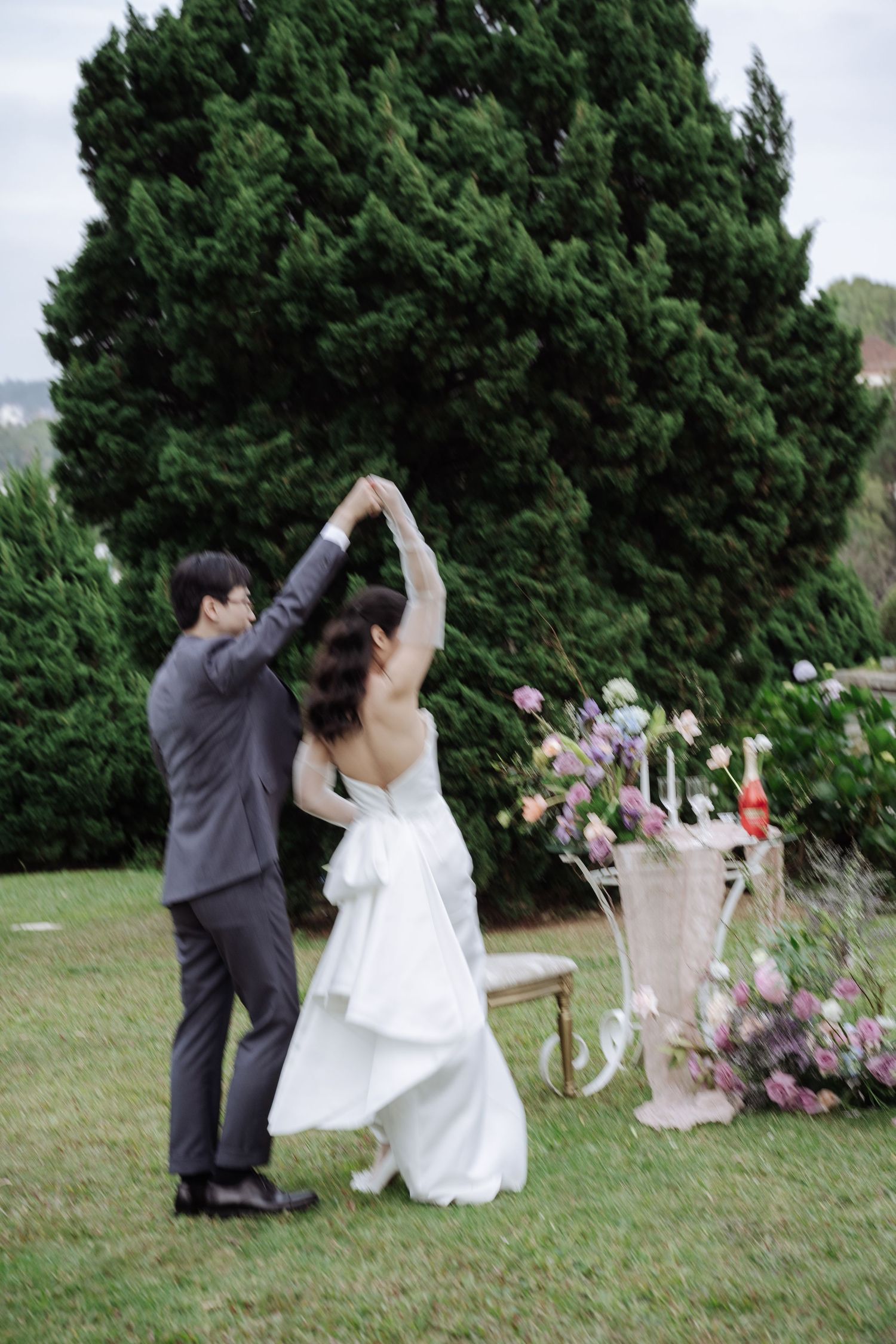 Bride and groom dancing outdoors near floral arrangements with lush green trees in background.