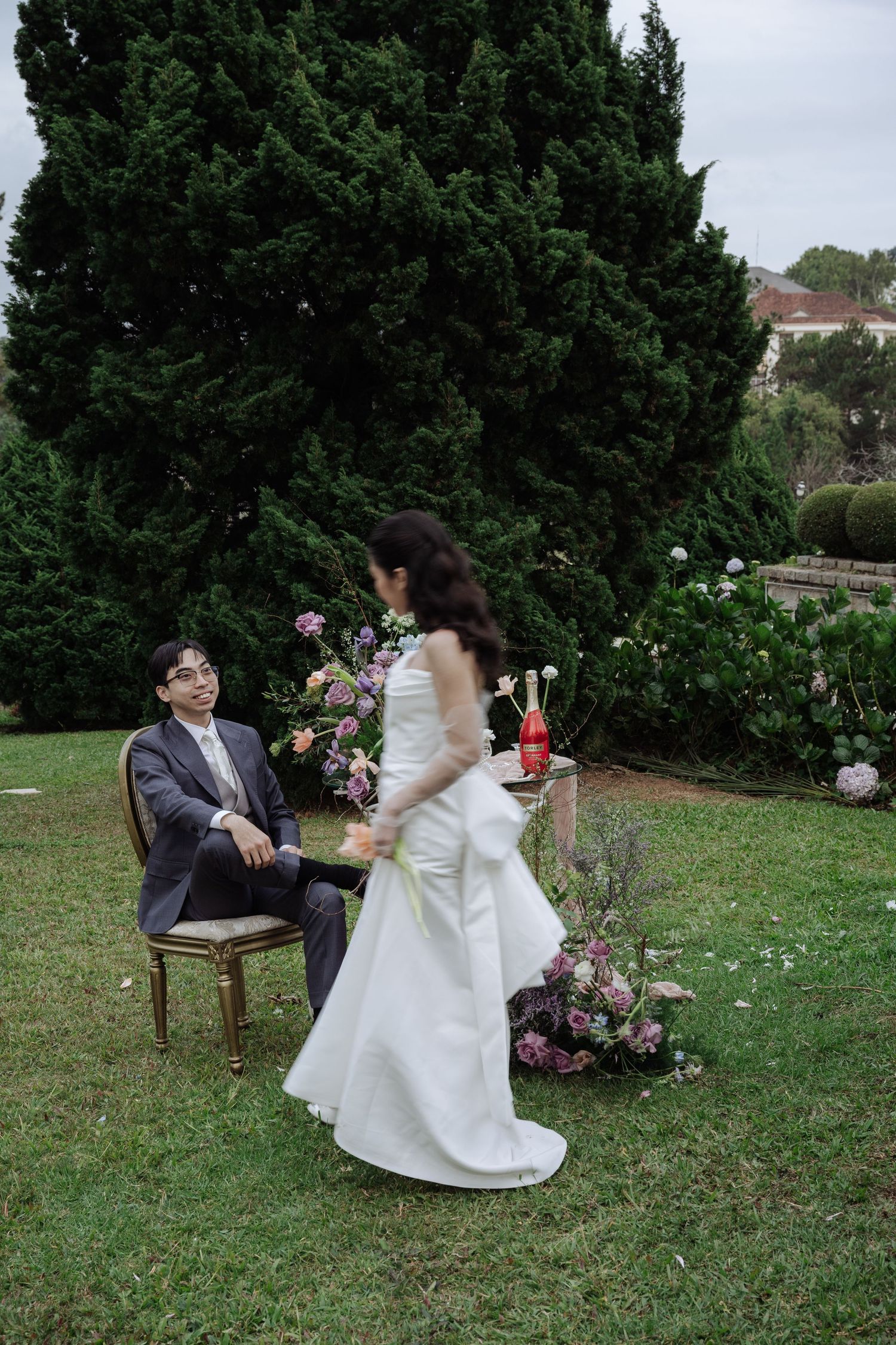 Groom seated in chair smiling at bride in white gown in lush garden wedding setting.