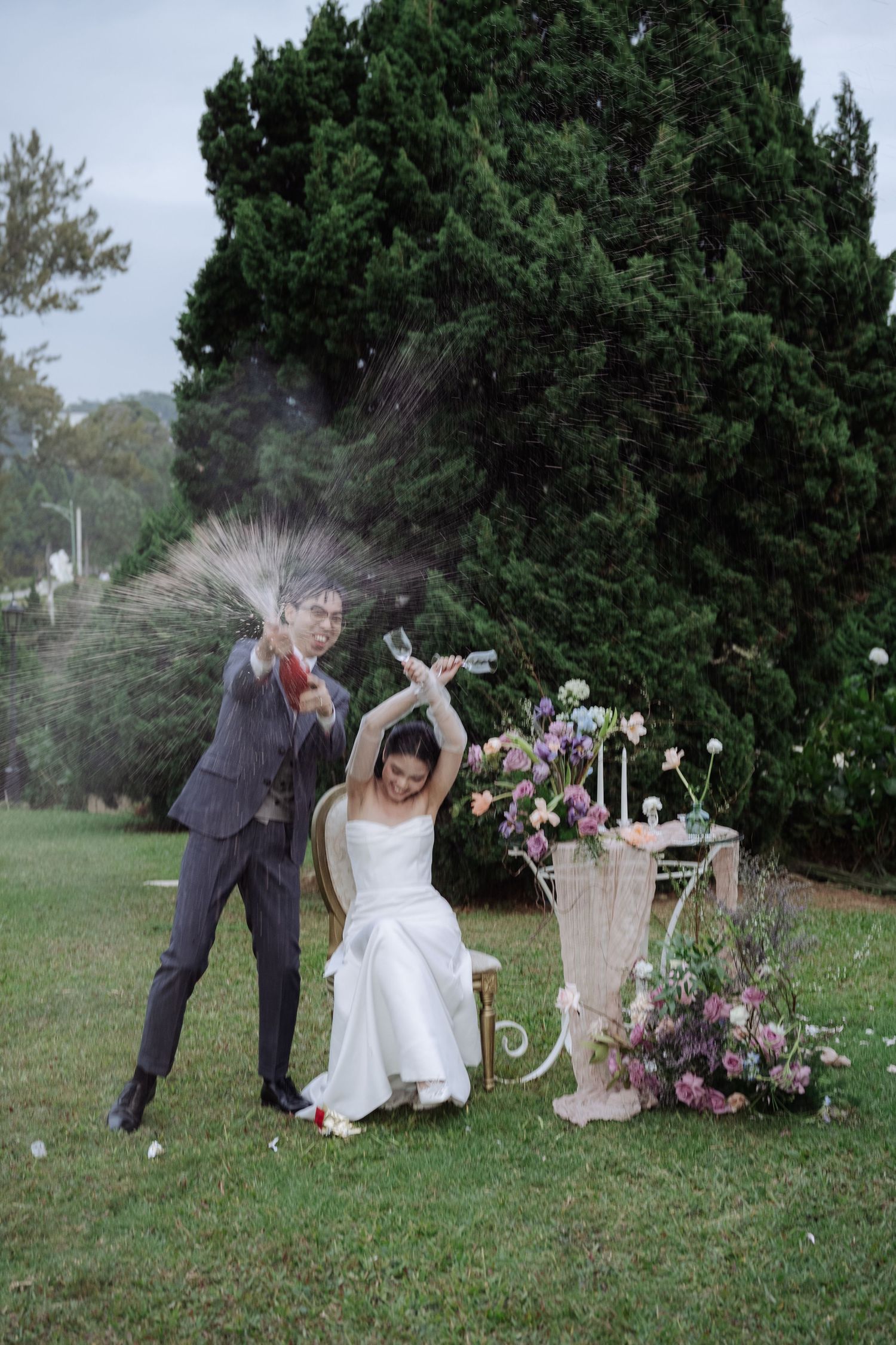 Joyful couple popping champagne bottle with spray at elegant outdoor garden wedding celebration.