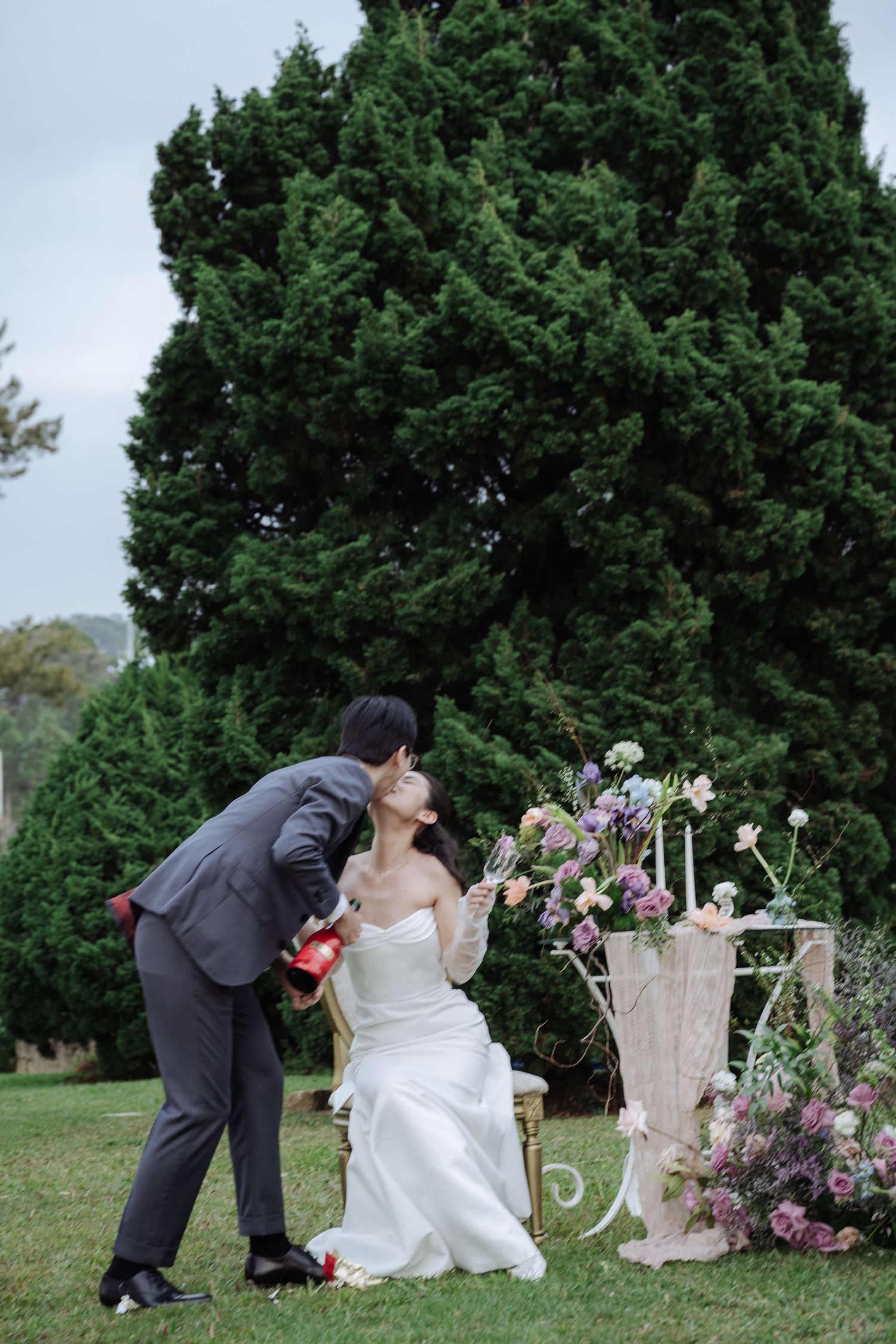 Groom leaning in to kiss seated bride in white gown beside colorful floral wedding decor.