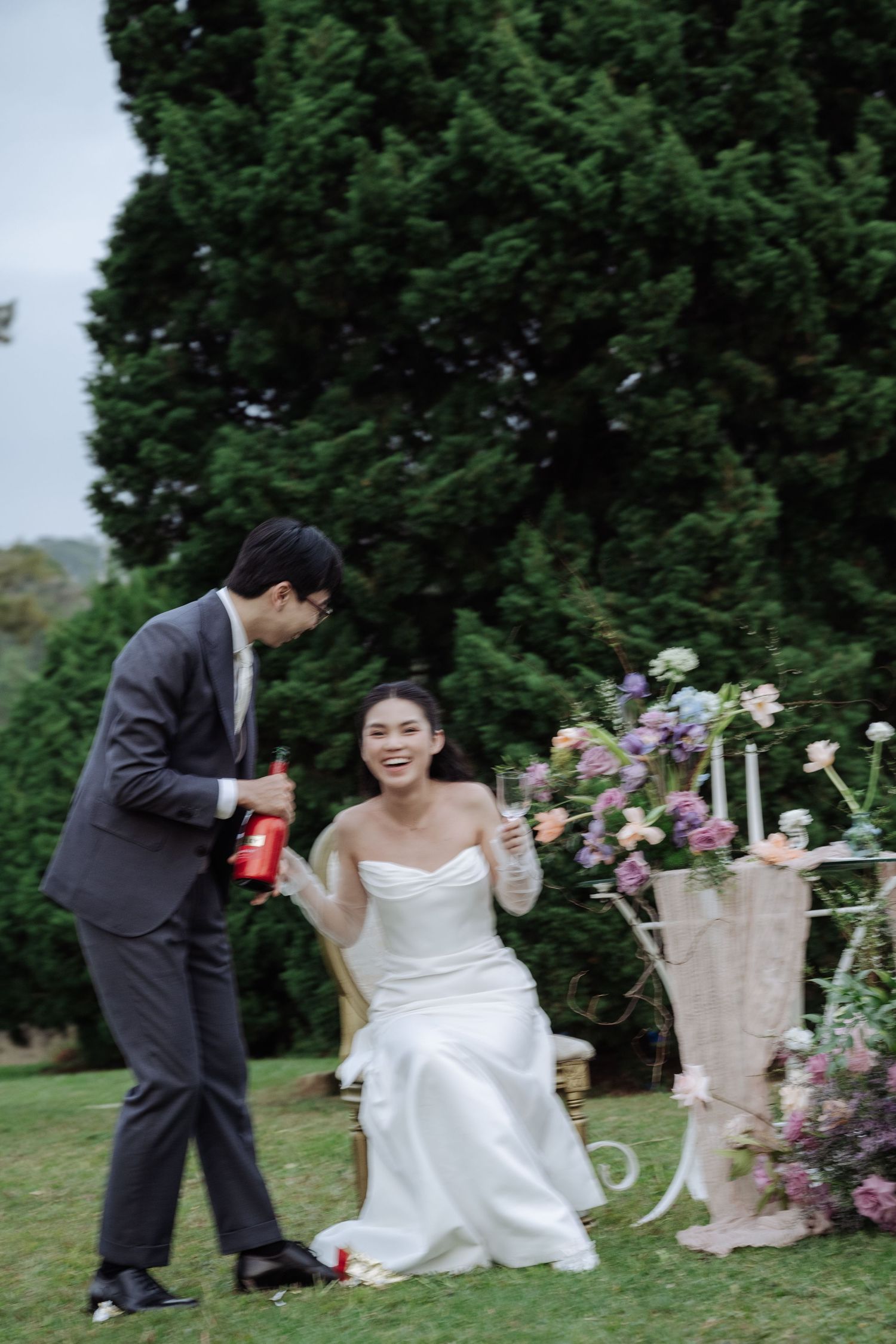 Smiling bride in white gown holding glass while groom pours champagne at garden wedding.