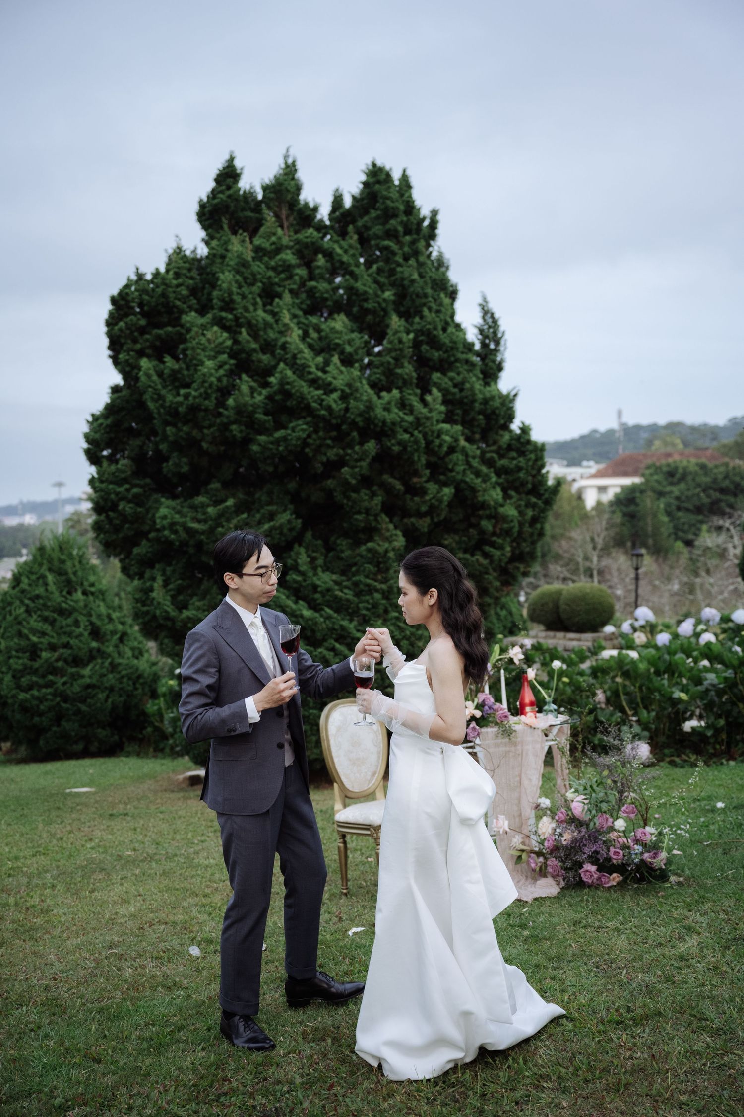 Bride and groom toast champagne glasses outdoors in a garden setting with floral arrangements nearby.