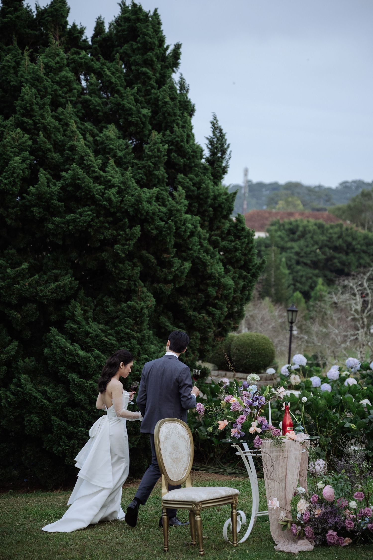 Bride and groom stand together near an elegant chair and floral display in a scenic garden.