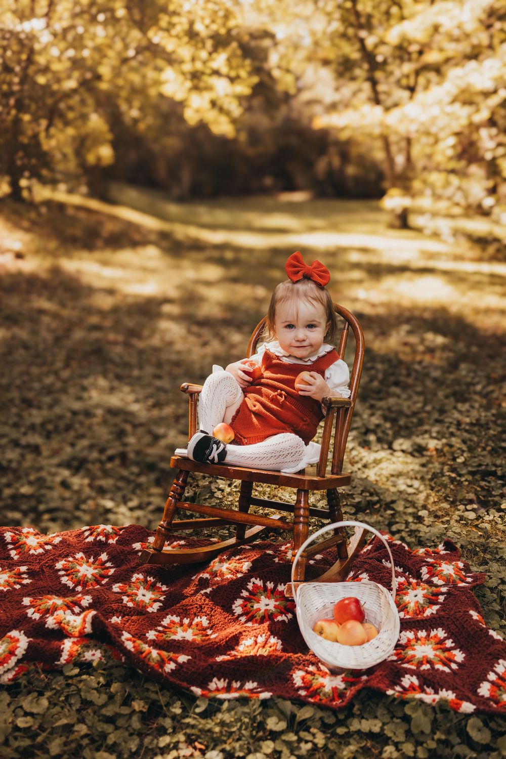 Toddler girl in red dress and bow sits in rocking chair on colorful blanket with apple basket in autumn park.