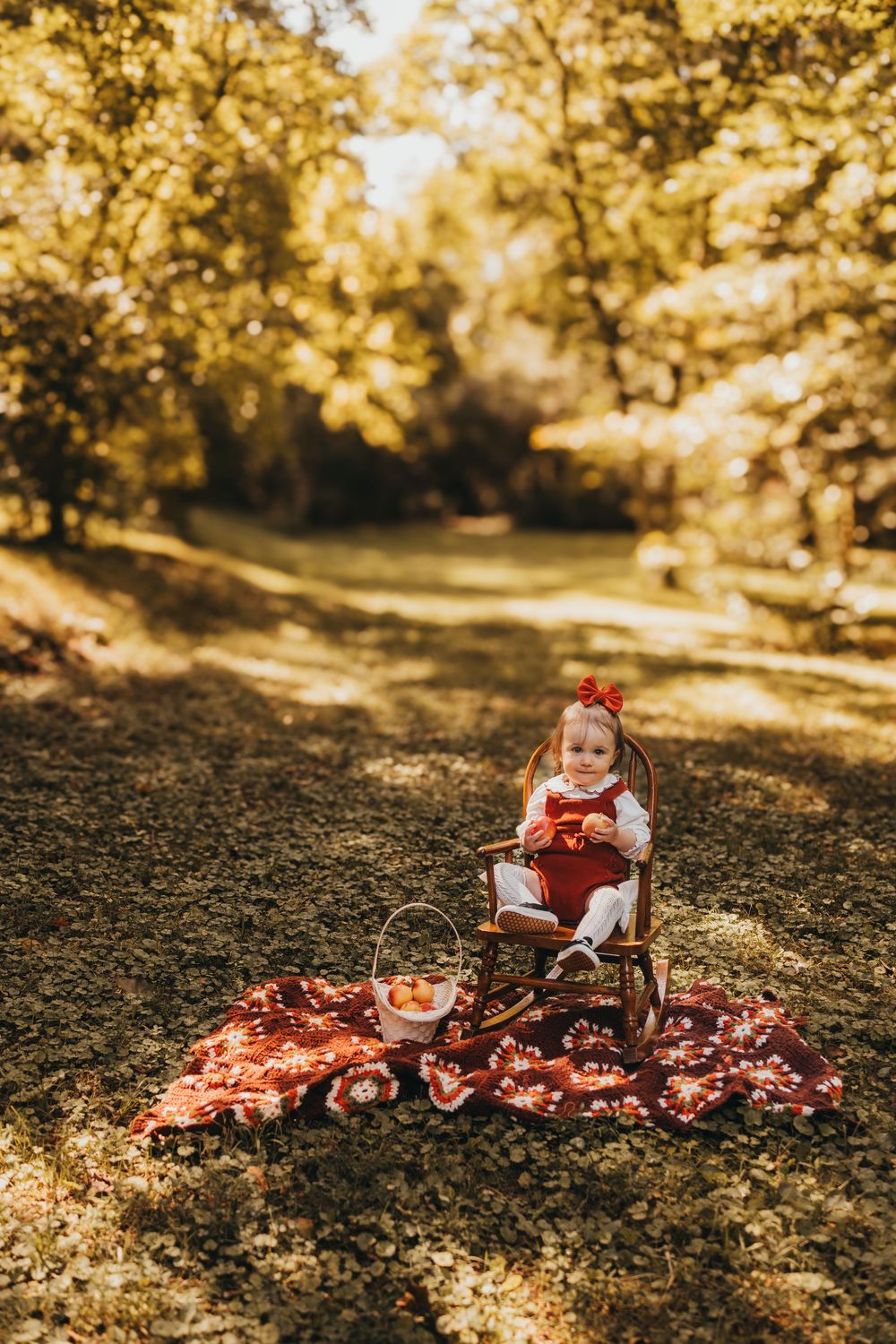 Adorable toddler girl in red dress with bow sits in wooden rocking chair on fall blanket outdoors.