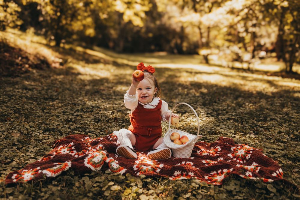 Baby girl in red romper sitting on floral blanket holding apple with basket of apples in sunny autumn park.