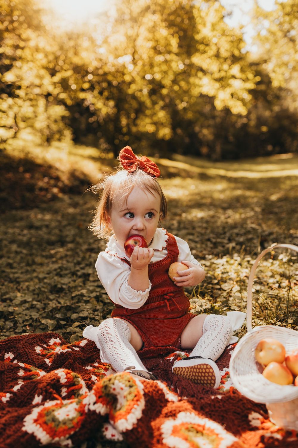 Cute toddler girl in red romper eating apple on colorful crochet blanket in golden autumn sunlight outdoors.
