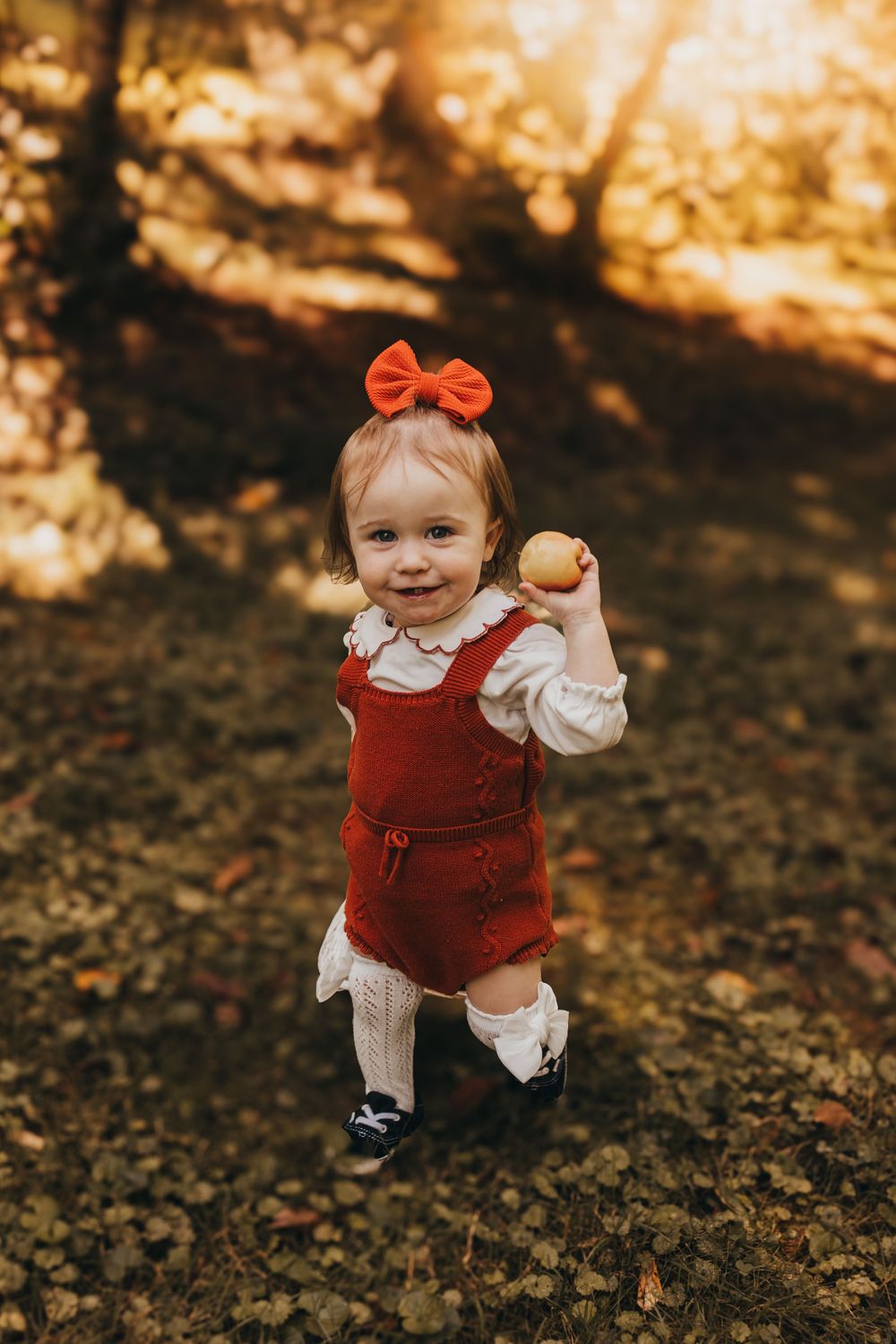 Happy toddler girl in red romper and bow holding apple while standing on fallen leaves in autumn park.
