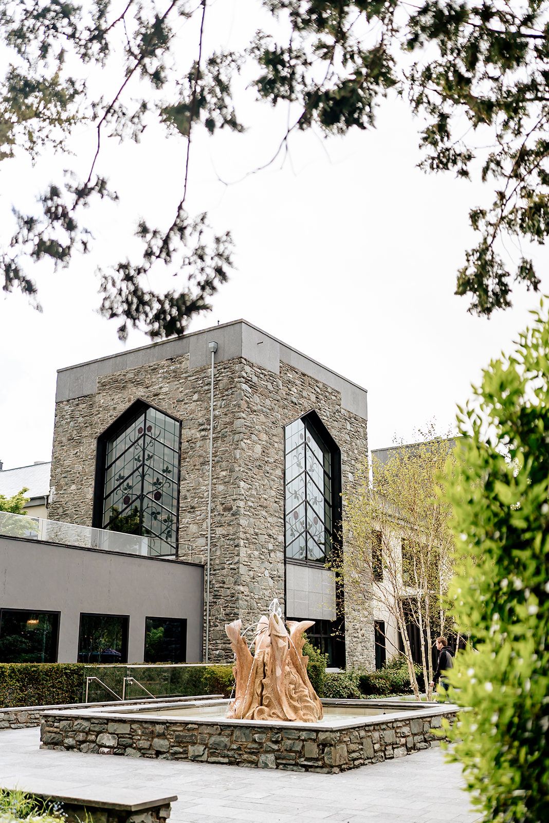 Modern stone building with large glass windows and angular architecture set among trees and landscaping.