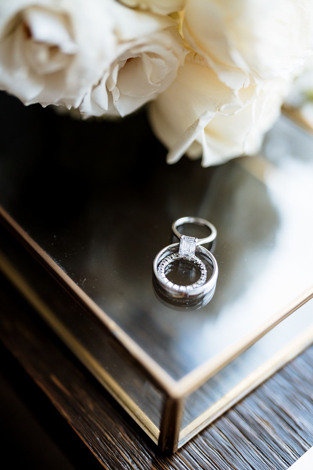 A pair of diamond wedding rings resting on a dark wooden surface beside white carnation flowers.