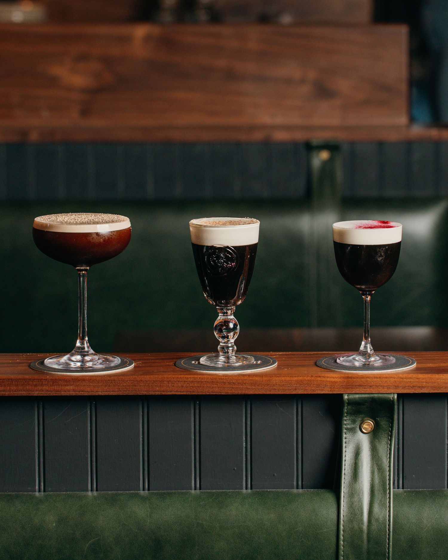 Different colored martini and cocktail glasses lined up on a dark wooden bar or shelf.