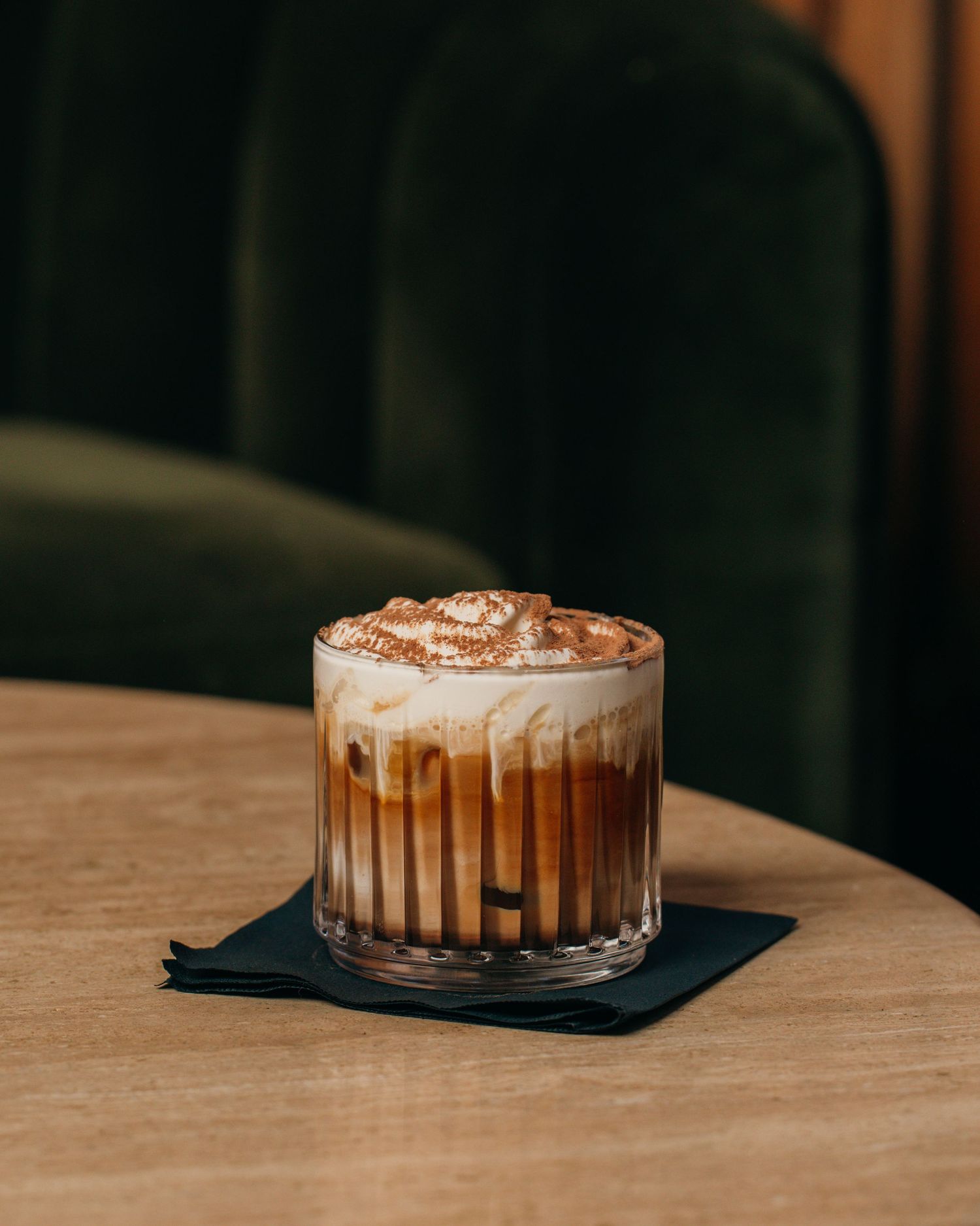 A glass of creamy iced coffee drink with layered caramel on wooden table against dark background.