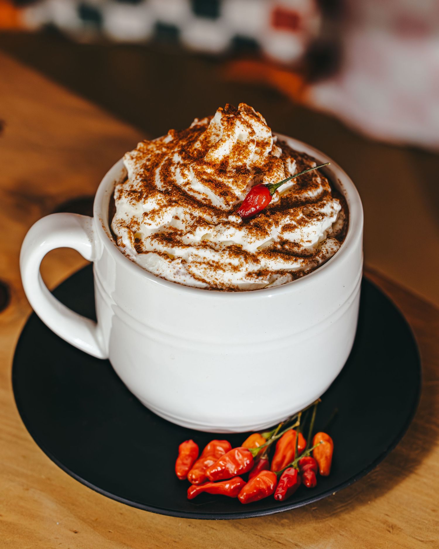 White coffee mug with whipped cream and cinnamon topping presented on a black plate on a wooden surface.