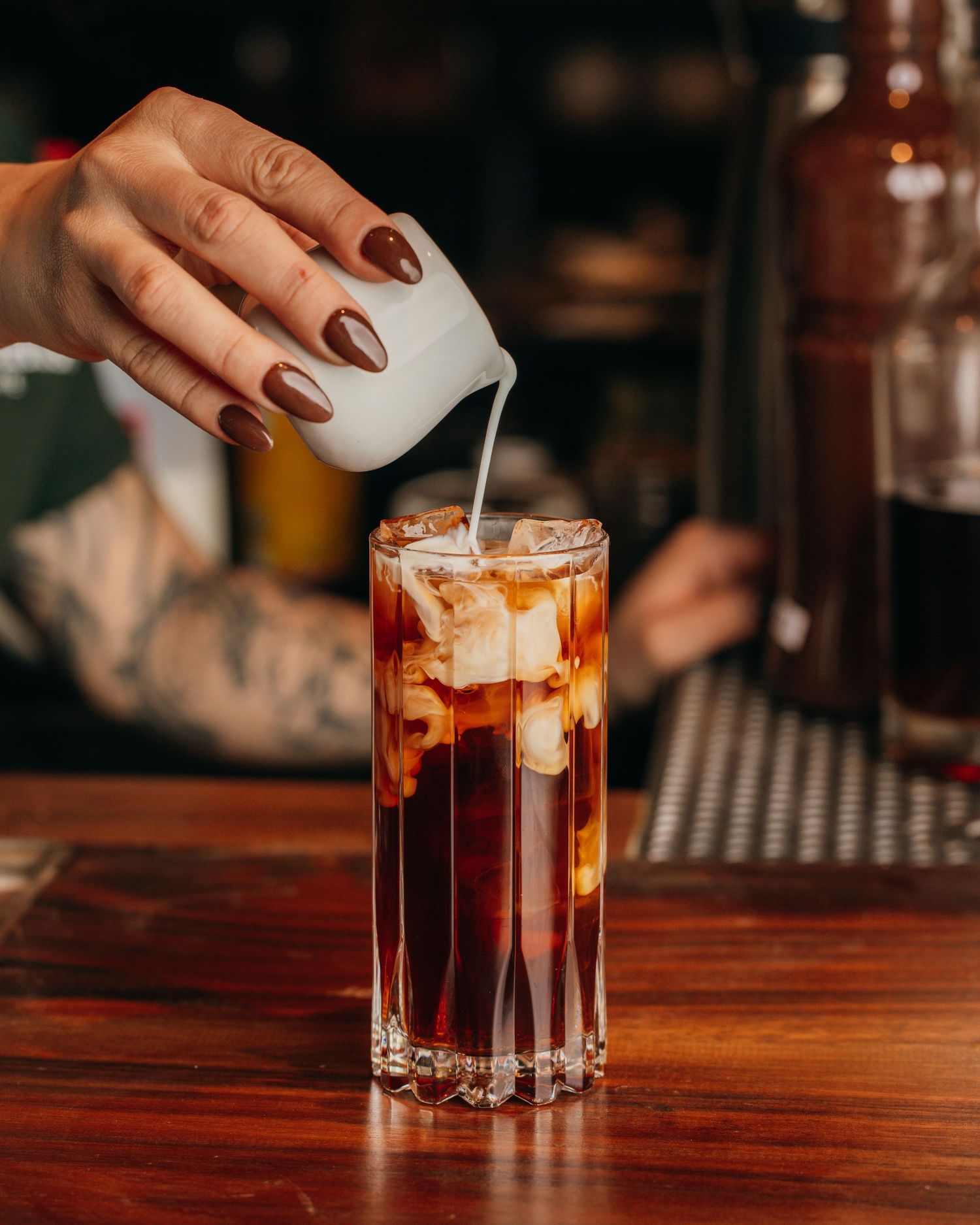 A hand dropping ice cubes into a layered cocktail at a dimly lit bar with rich wooden surfaces.