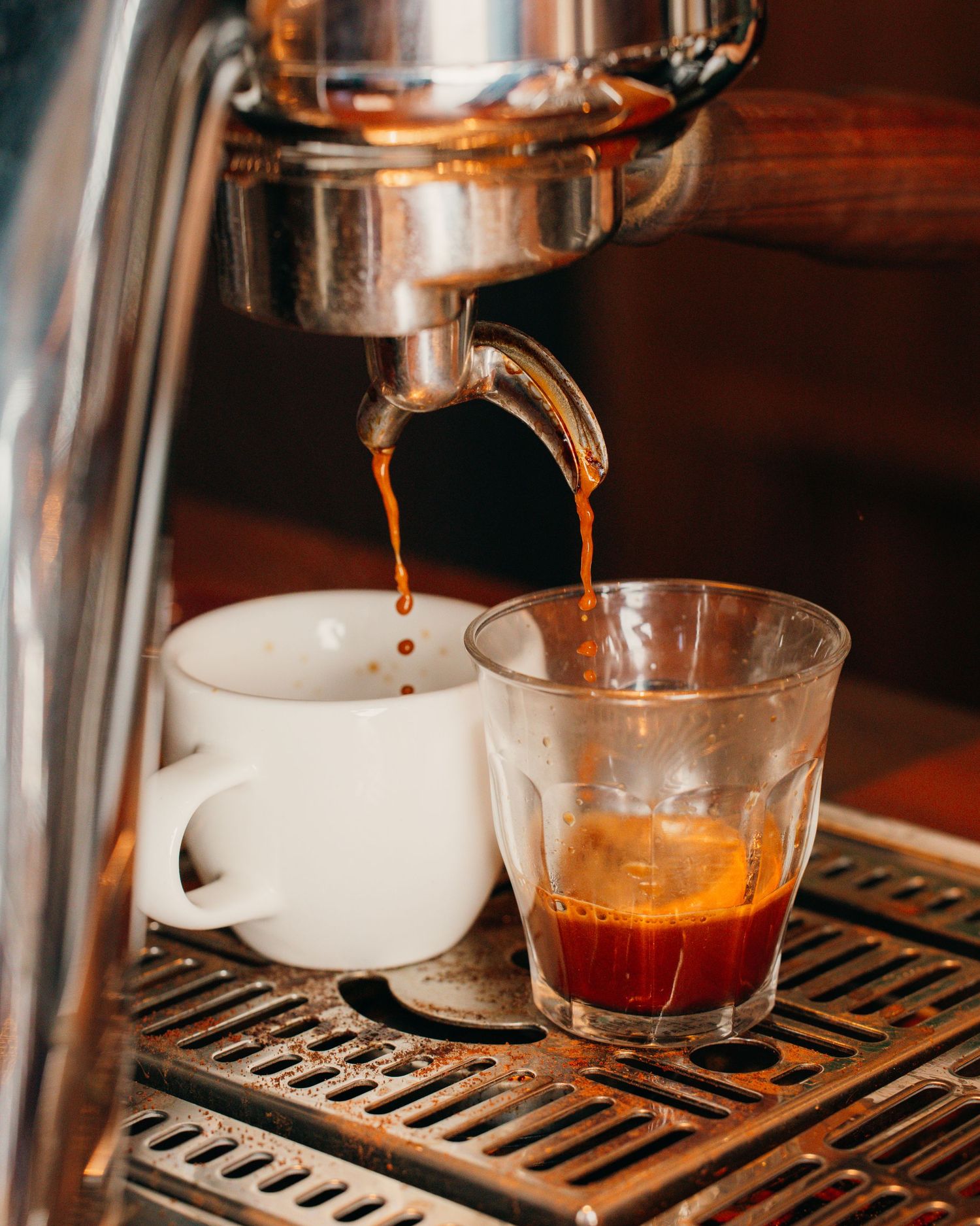 Fresh espresso shot pouring from a commercial coffee machine into a glass cup on a metal drip tray.