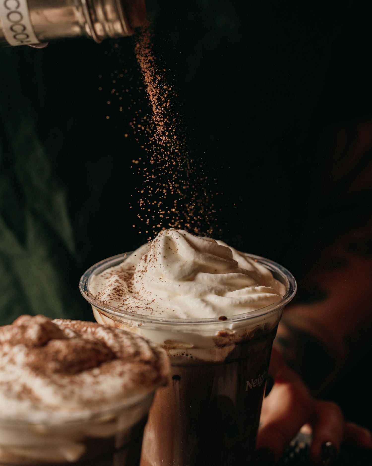 Dark moody photo of cocoa powder being dusted over whipped cream on a beverage in dramatic lighting.
