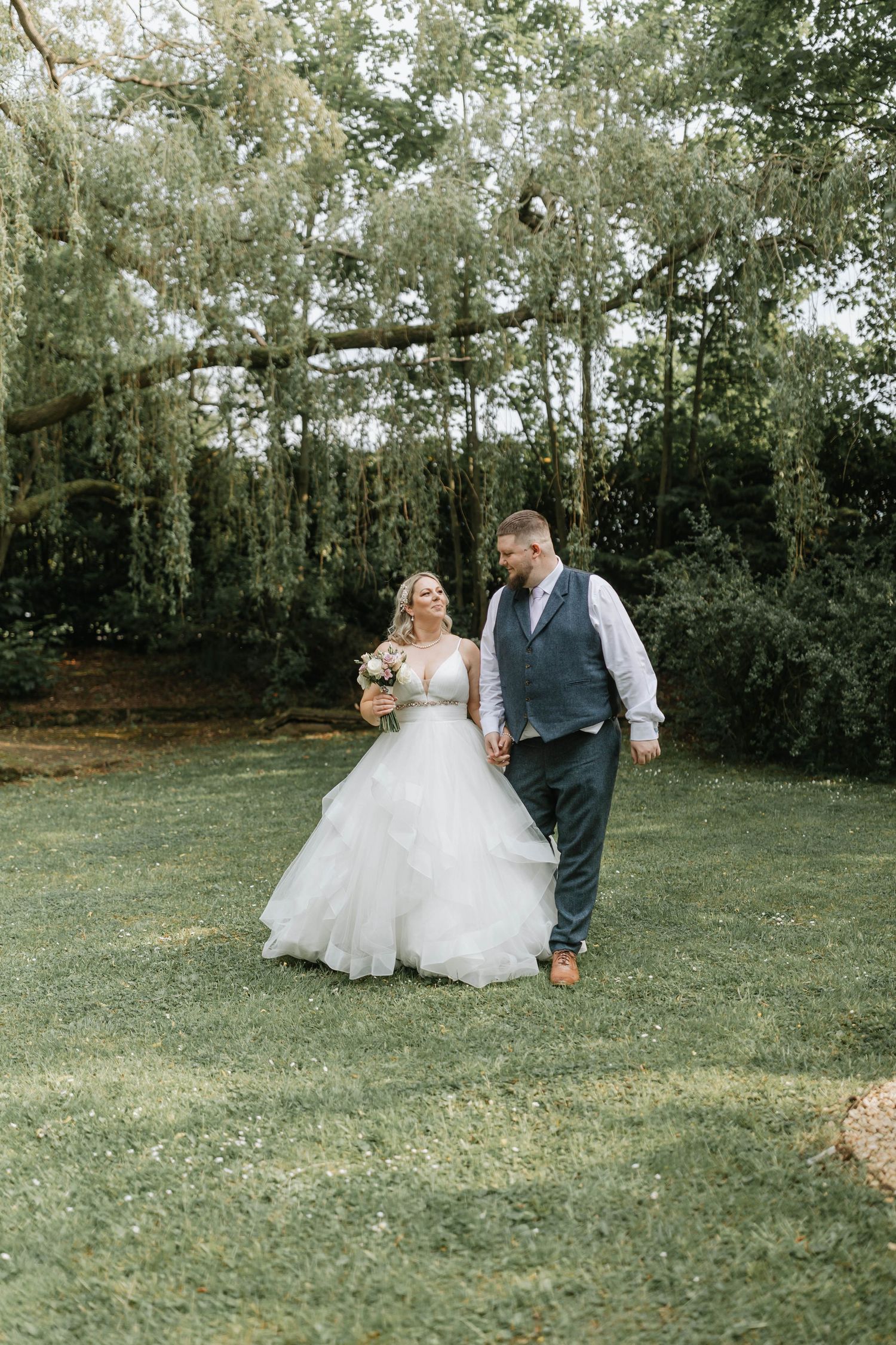 A bride in a white ballgown and groom in a vest take photos in a serene outdoor garden setting with lush trees.