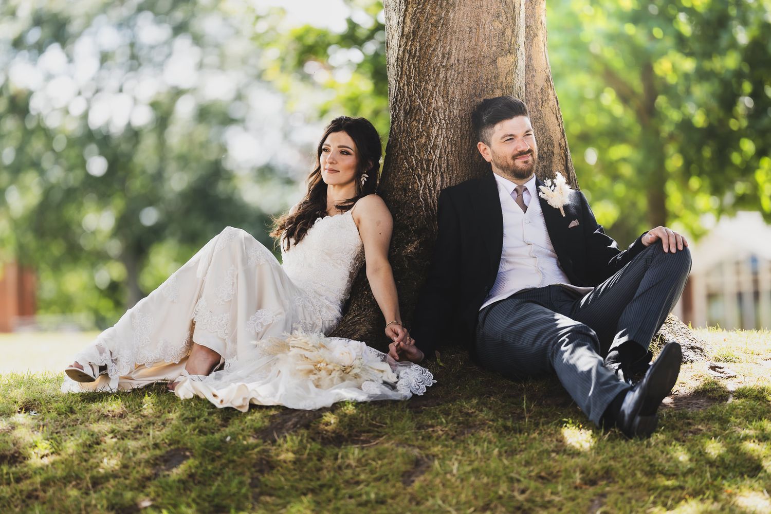A couple in wedding attire sits together under a large tree in a scenic outdoor setting during their wedding photoshoot.