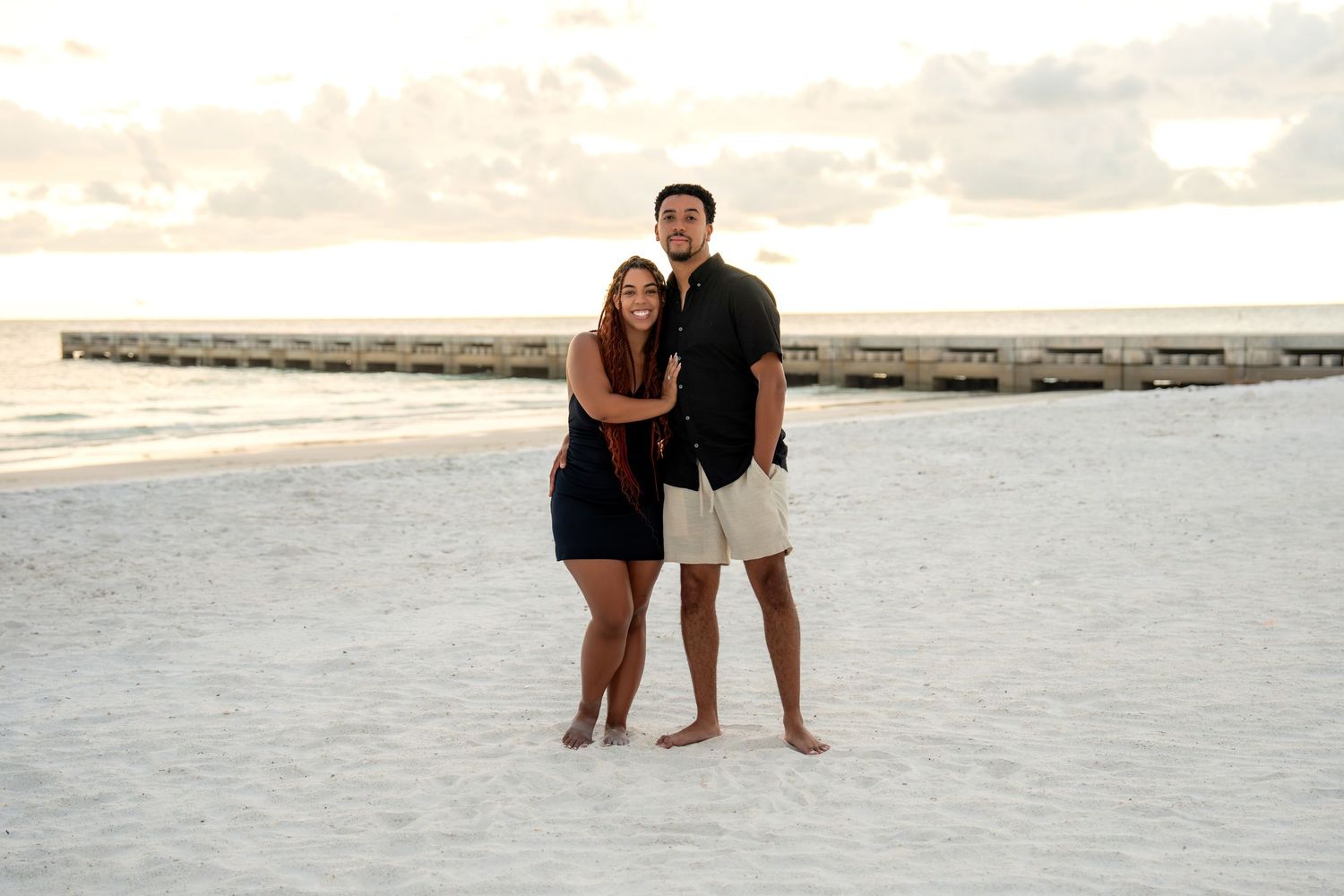 Smiling couple embracing on a white sandy beach with a pier in the background at sunset.