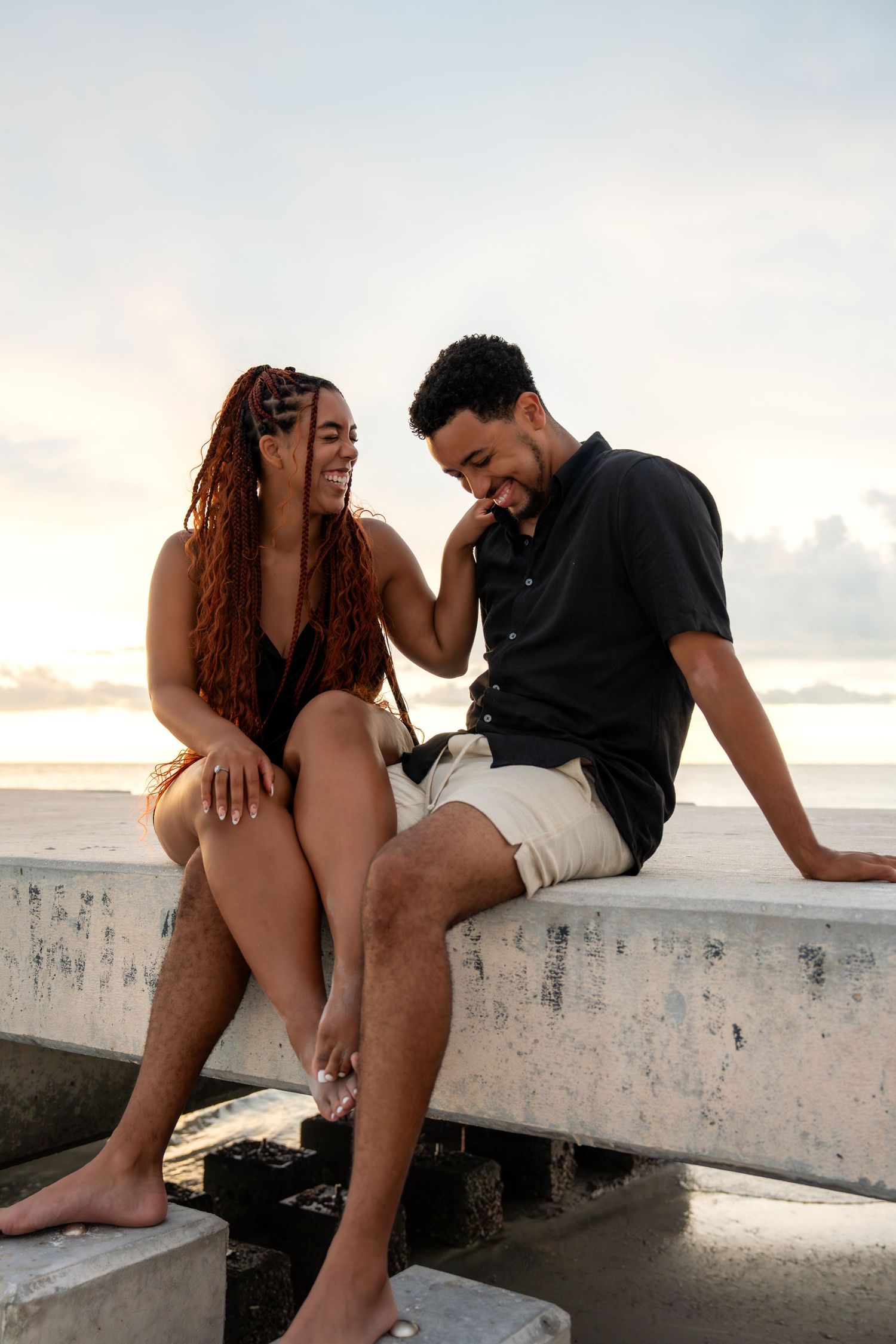 Laughing couple sitting together on a concrete pier at the beach during golden hour sunset.