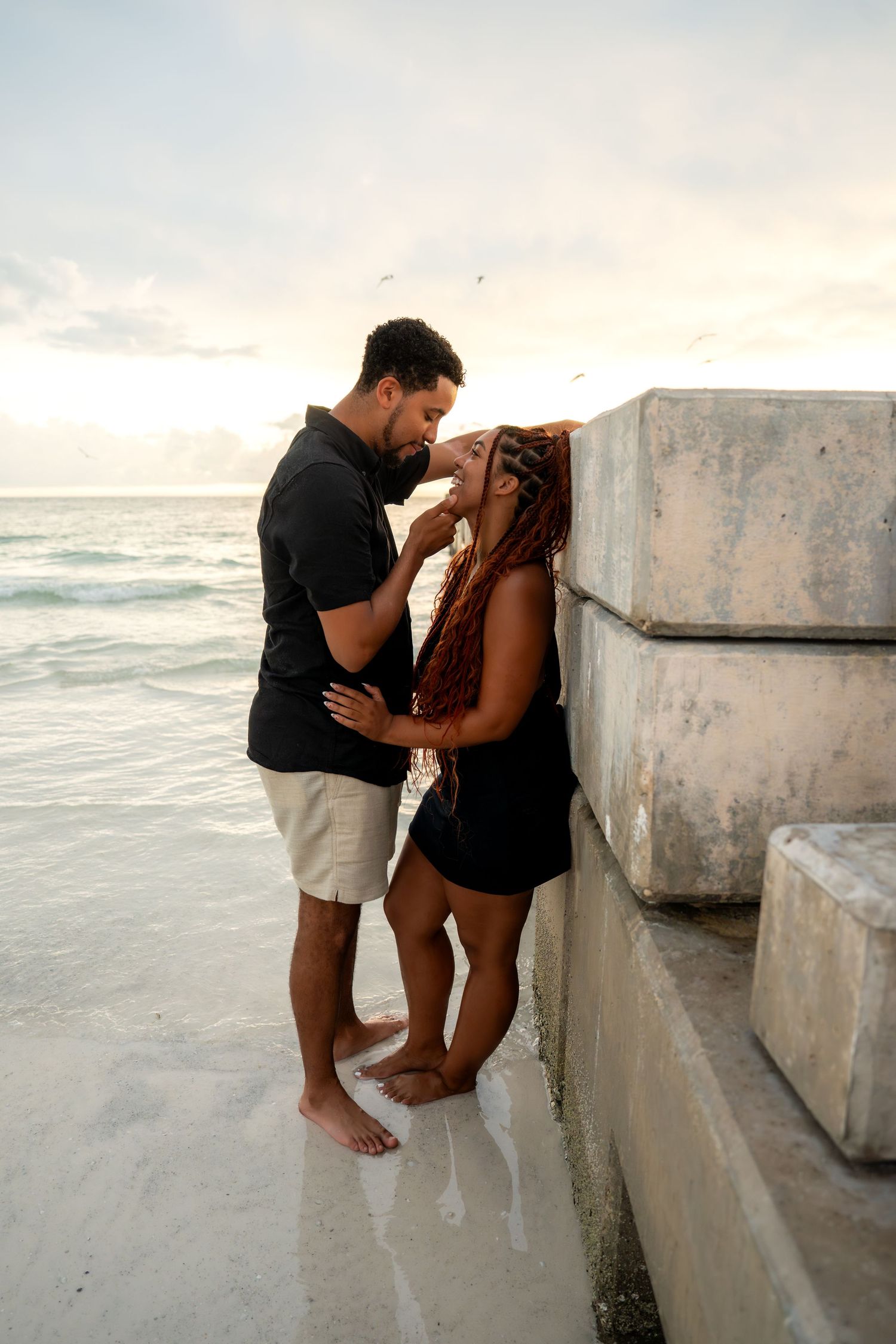 Couple sharing an intimate moment leaning against concrete pier blocks on a beach at sunset.