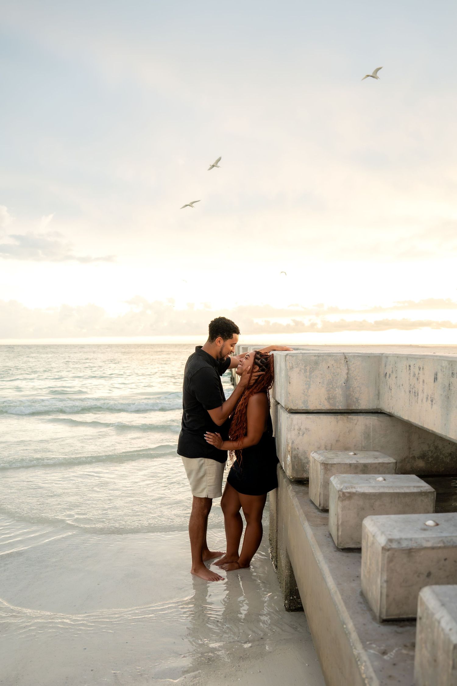 Couple sharing a romantic moment by concrete pier blocks on beach with seagulls at sunset.