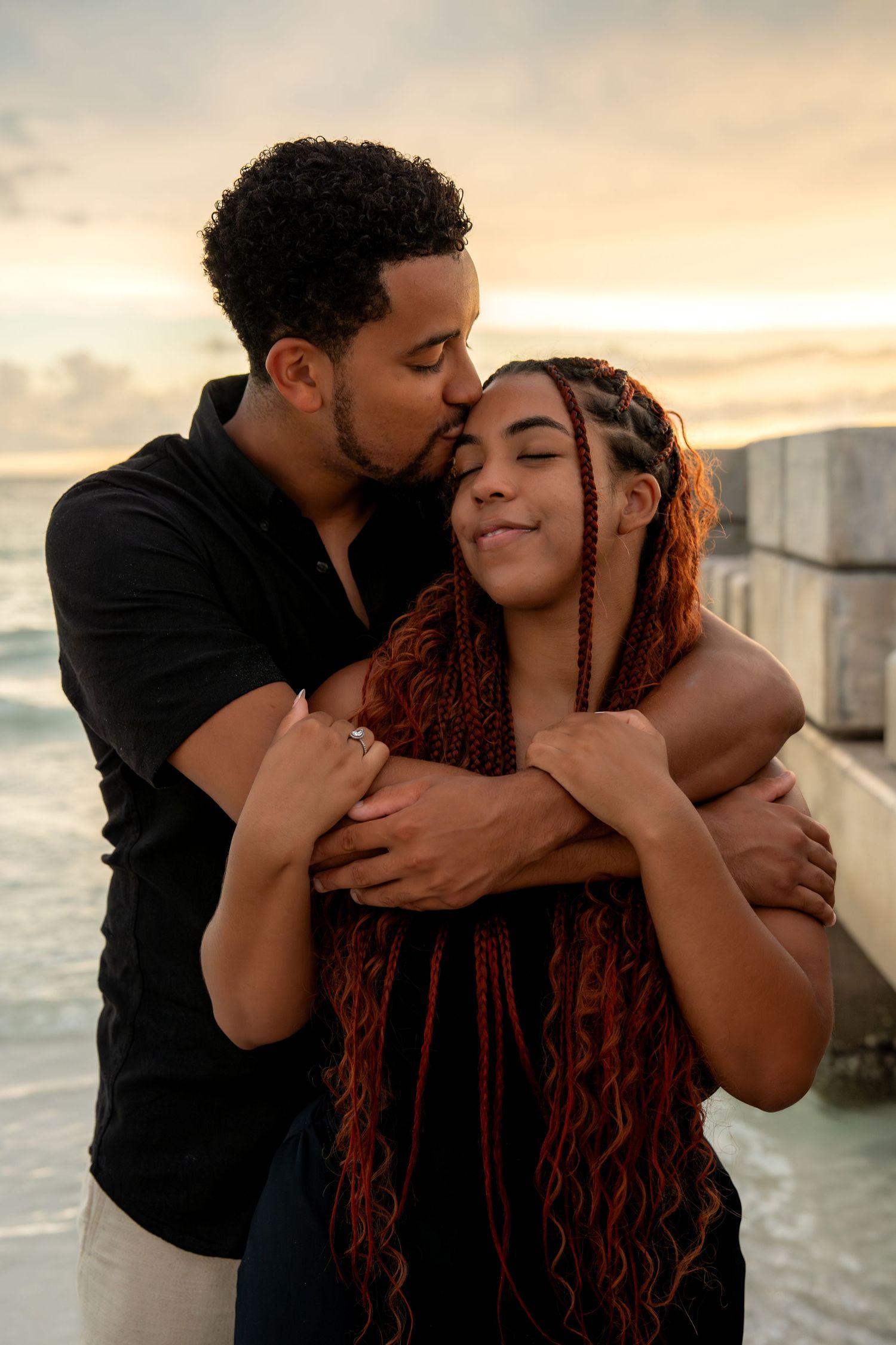 Man tenderly kissing woman on forehead while embracing her from behind at beach sunset.