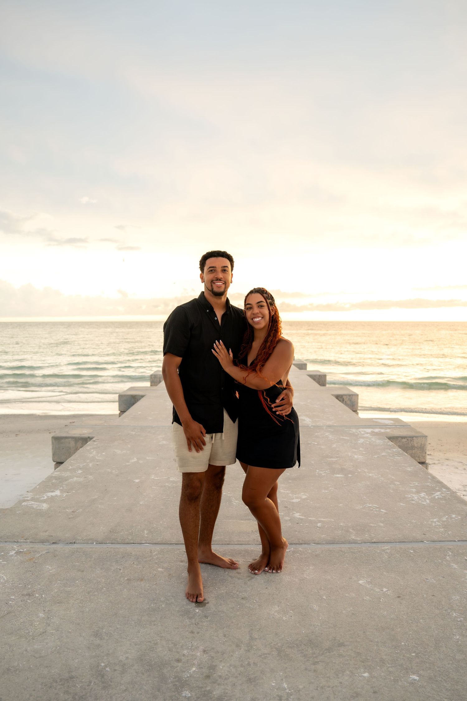 Smiling couple standing together on concrete pier with beautiful ocean sunset backdrop.